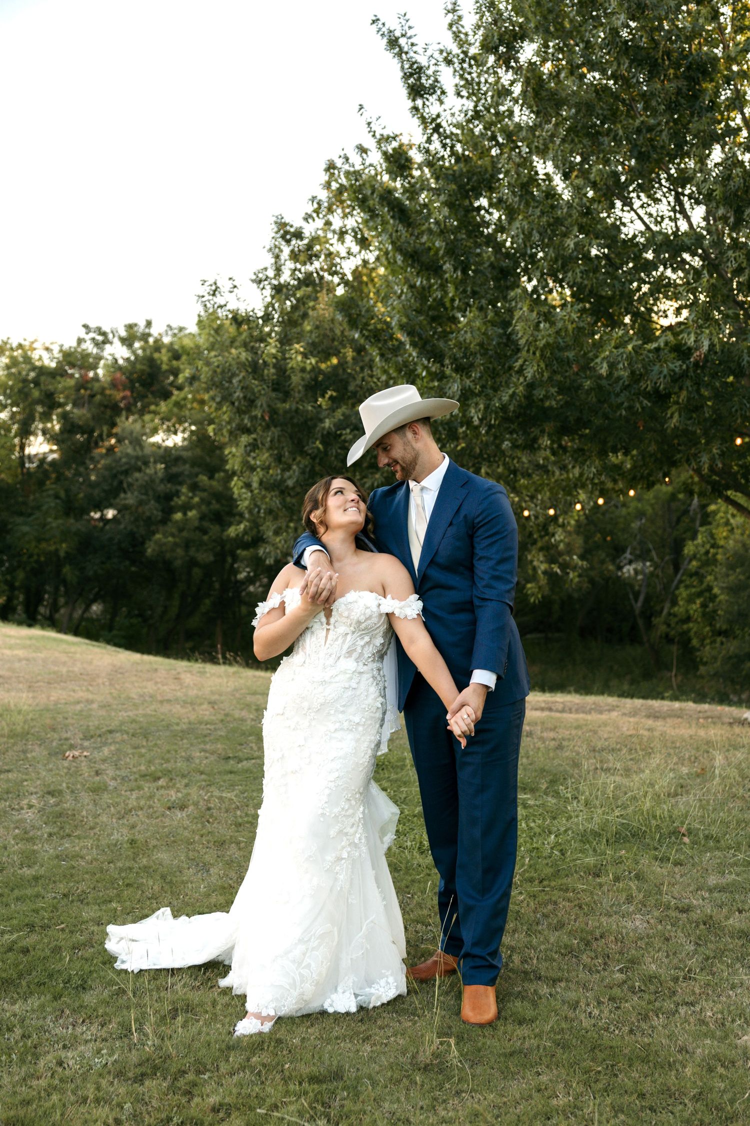 Bride in floral gown and groom in navy suit with cowboy hat holding hands in green meadow.