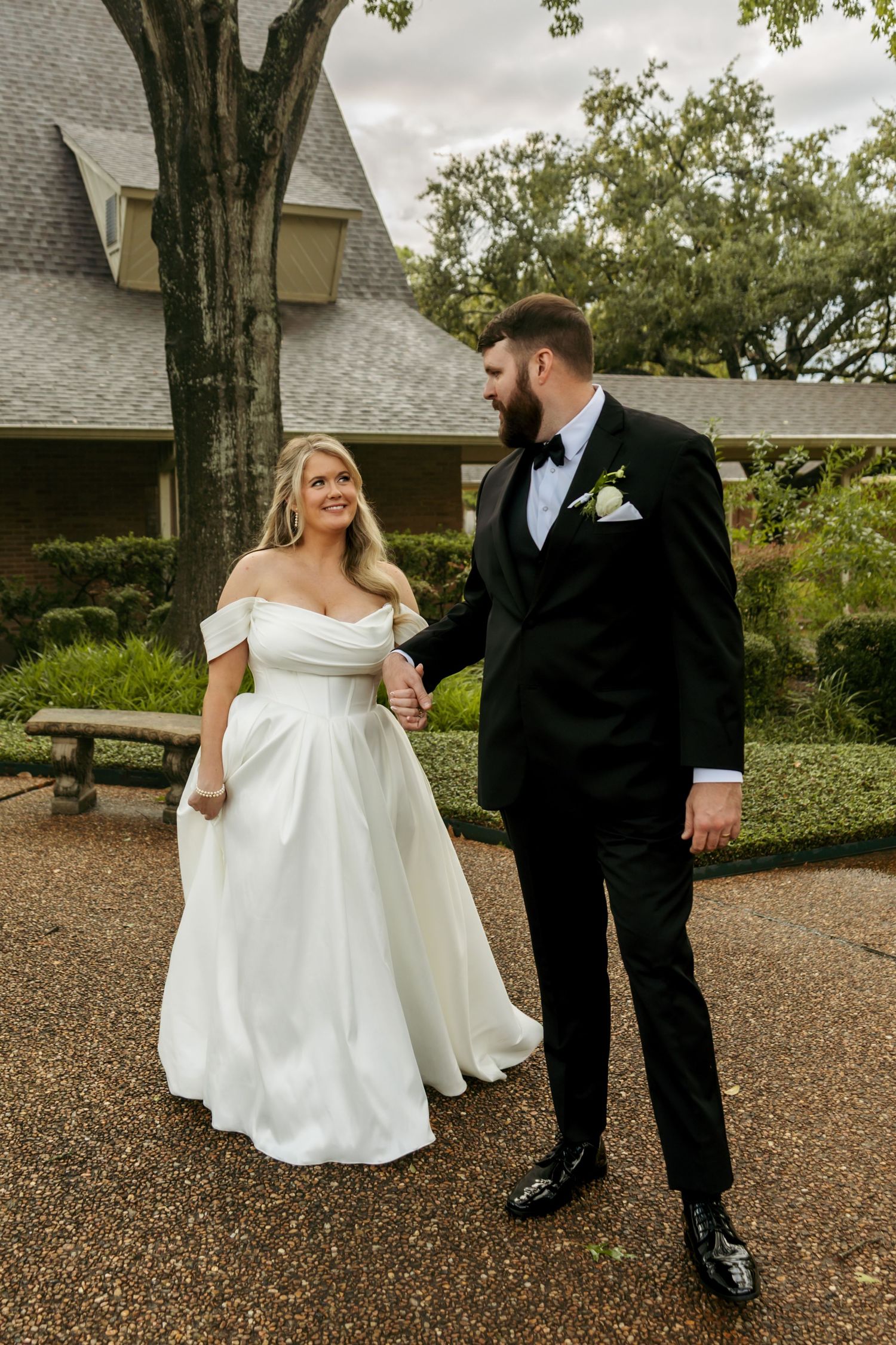 Smiling bride in off-shoulder gown walks hand-in-hand with bearded groom in black tuxedo at outdoor wedding.