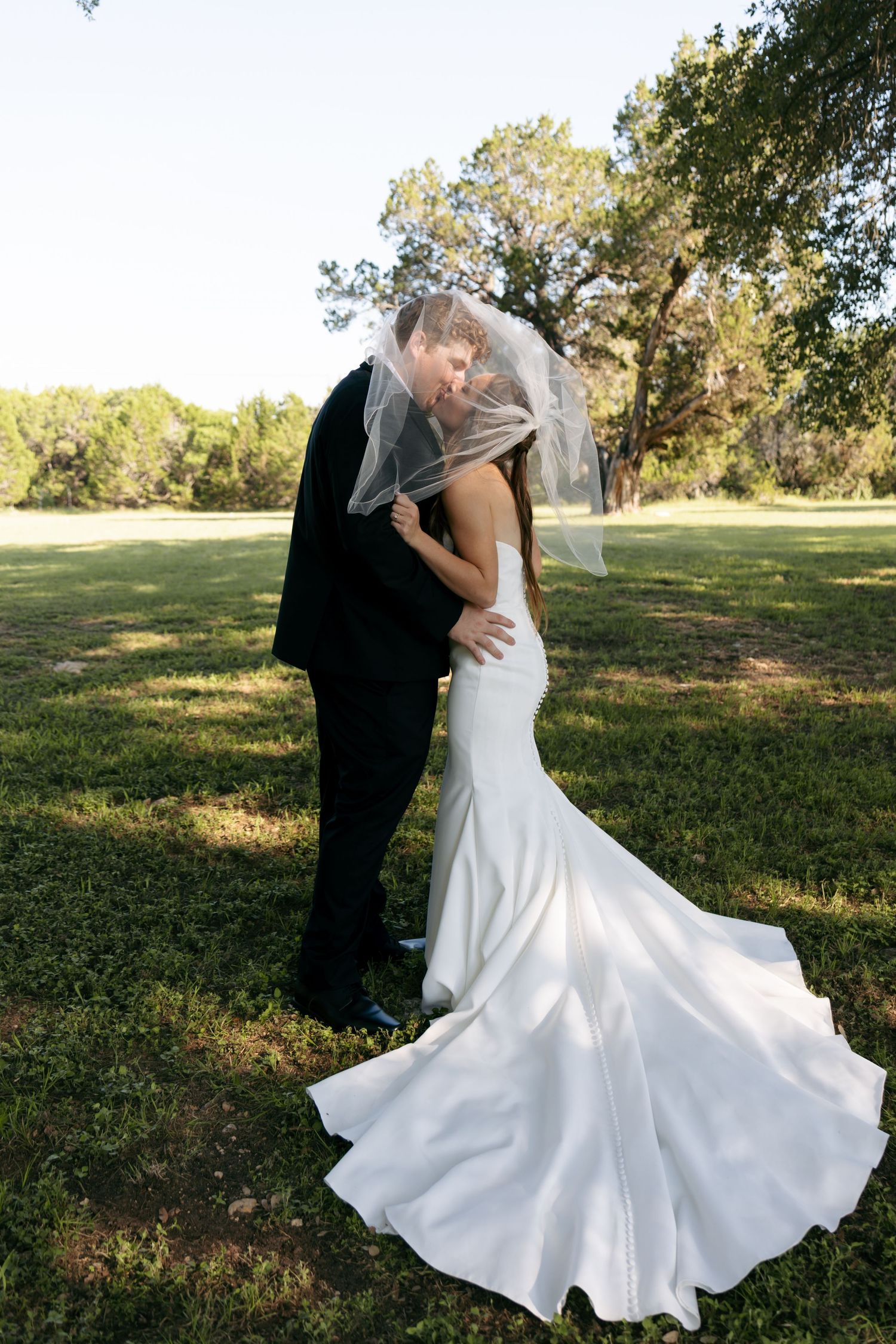 A bride and groom share a kiss under a flowing veil on a green lawn surrounded by trees.
