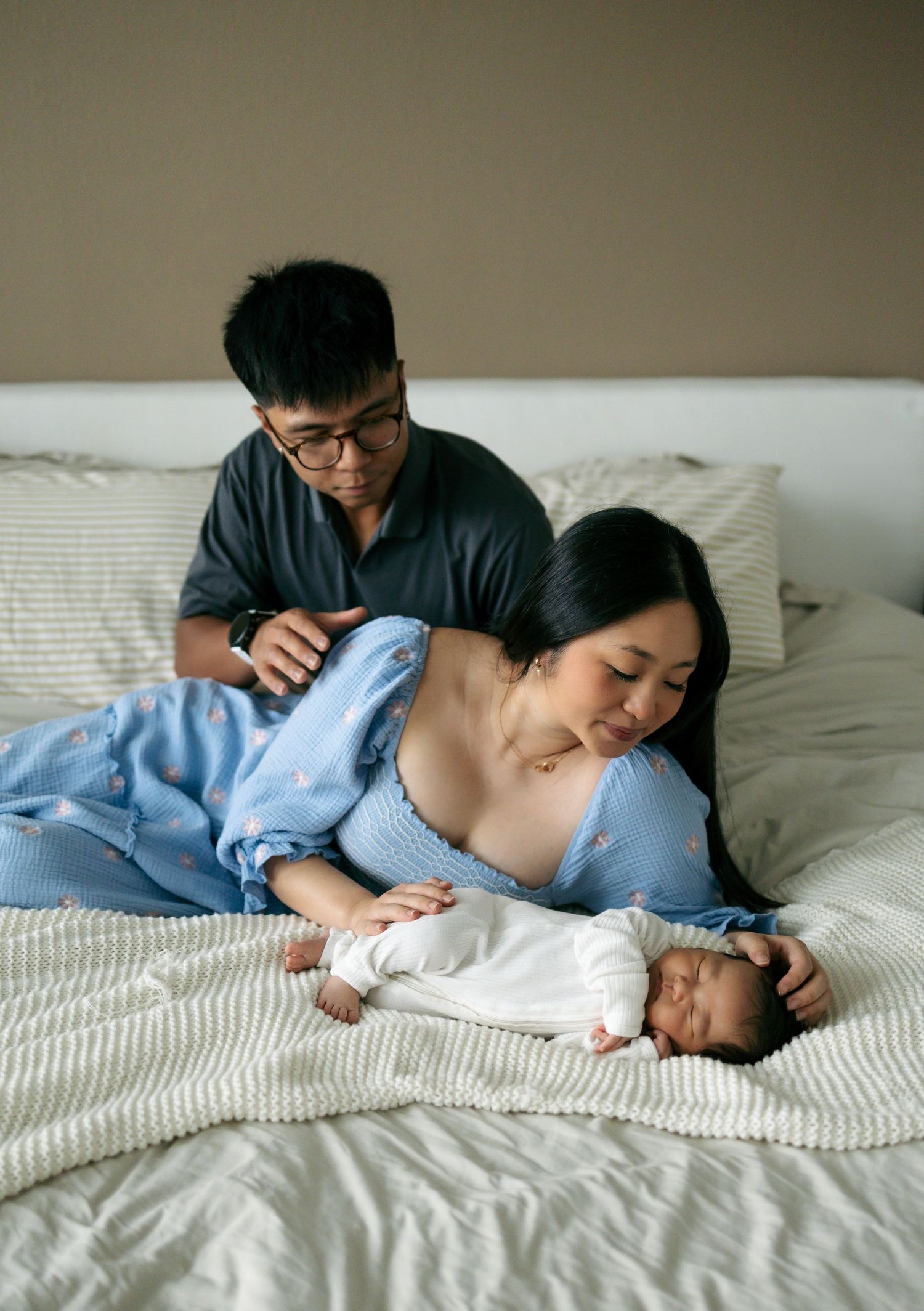 Asian parents tenderly watch over their sleeping newborn baby on a white bed.