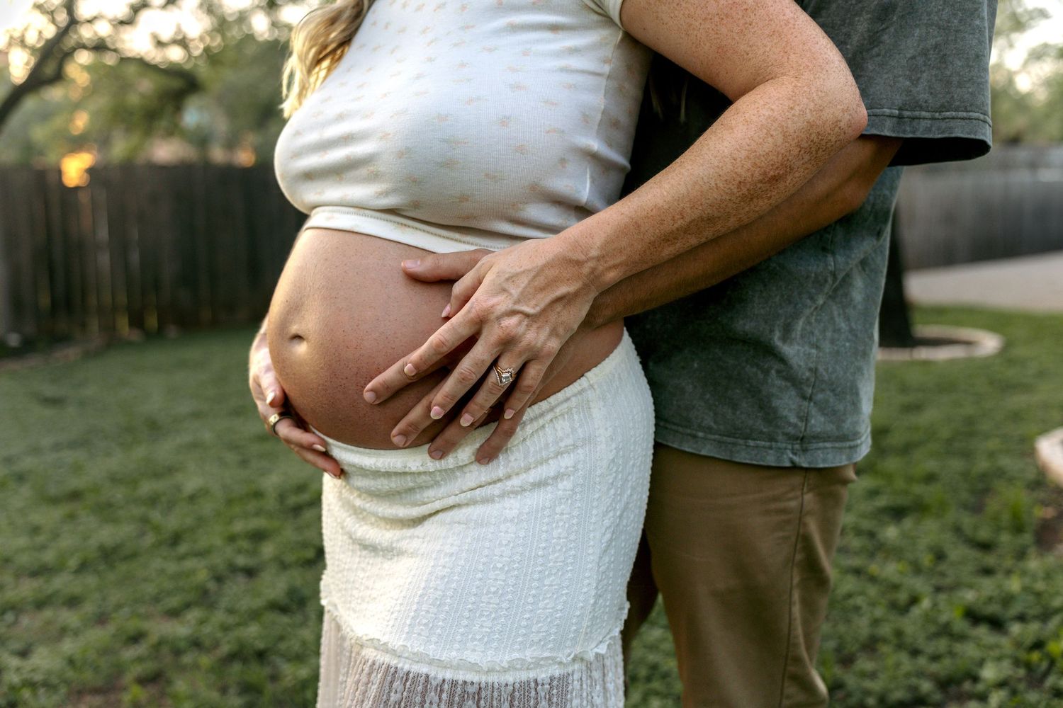 Close-up of pregnant belly with partner embracing woman in white dress outdoors.