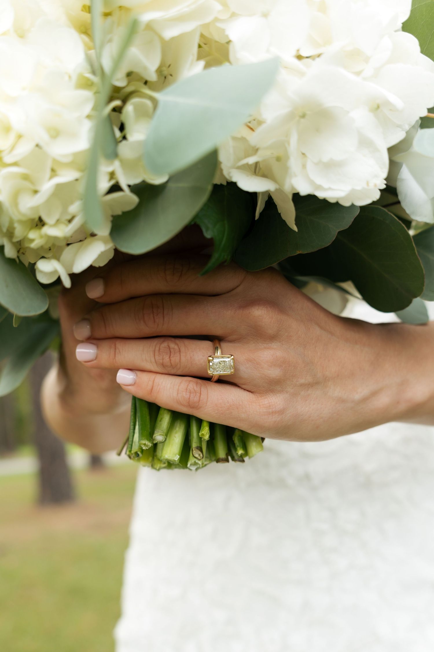 Bride holding white hydrangea bouquet with green stems, showcasing elegant emerald-cut diamond engagement ring on gold band.