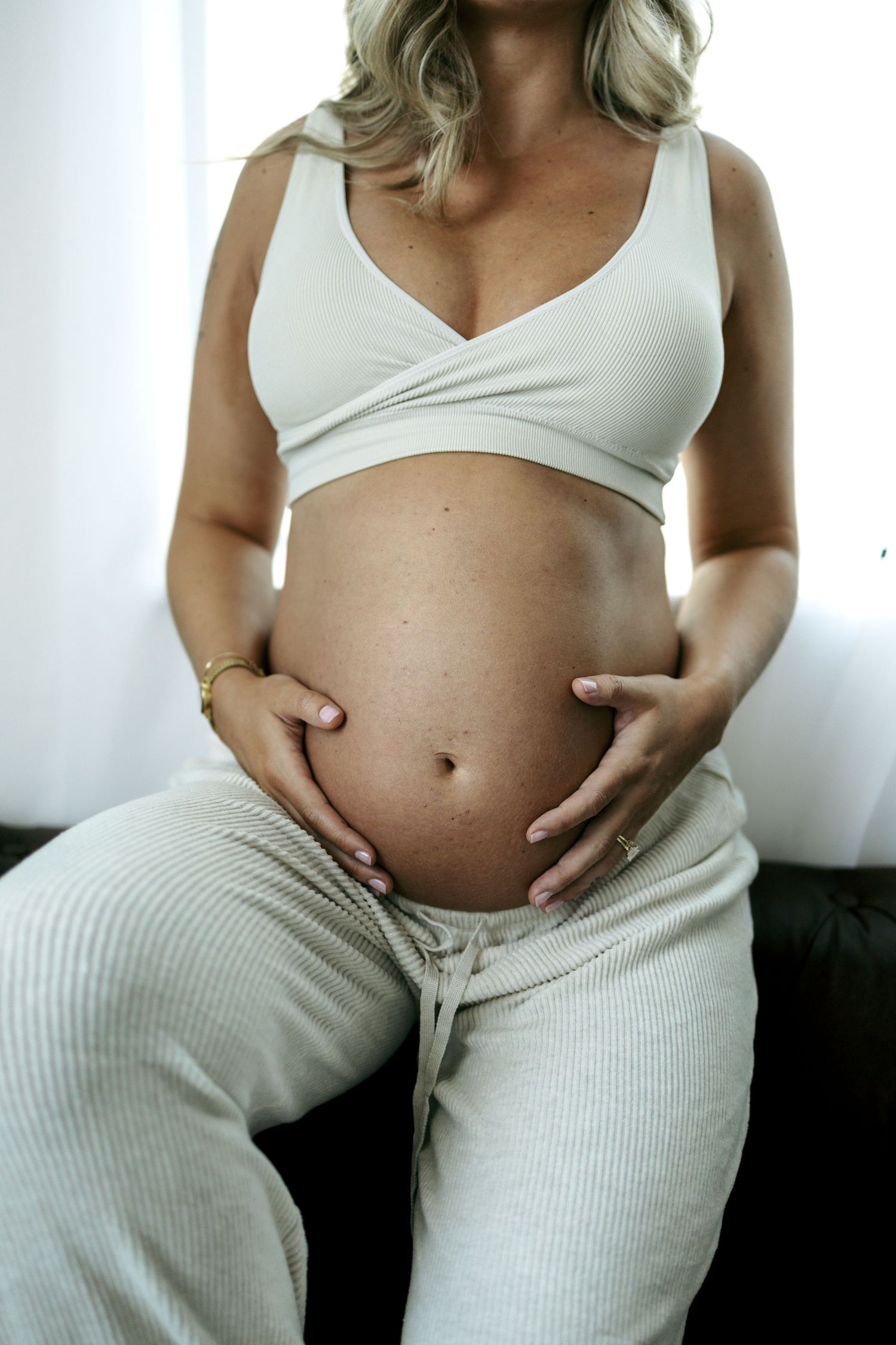 Pregnant woman in white crop top and ribbed pants cradling her baby bump with both hands.