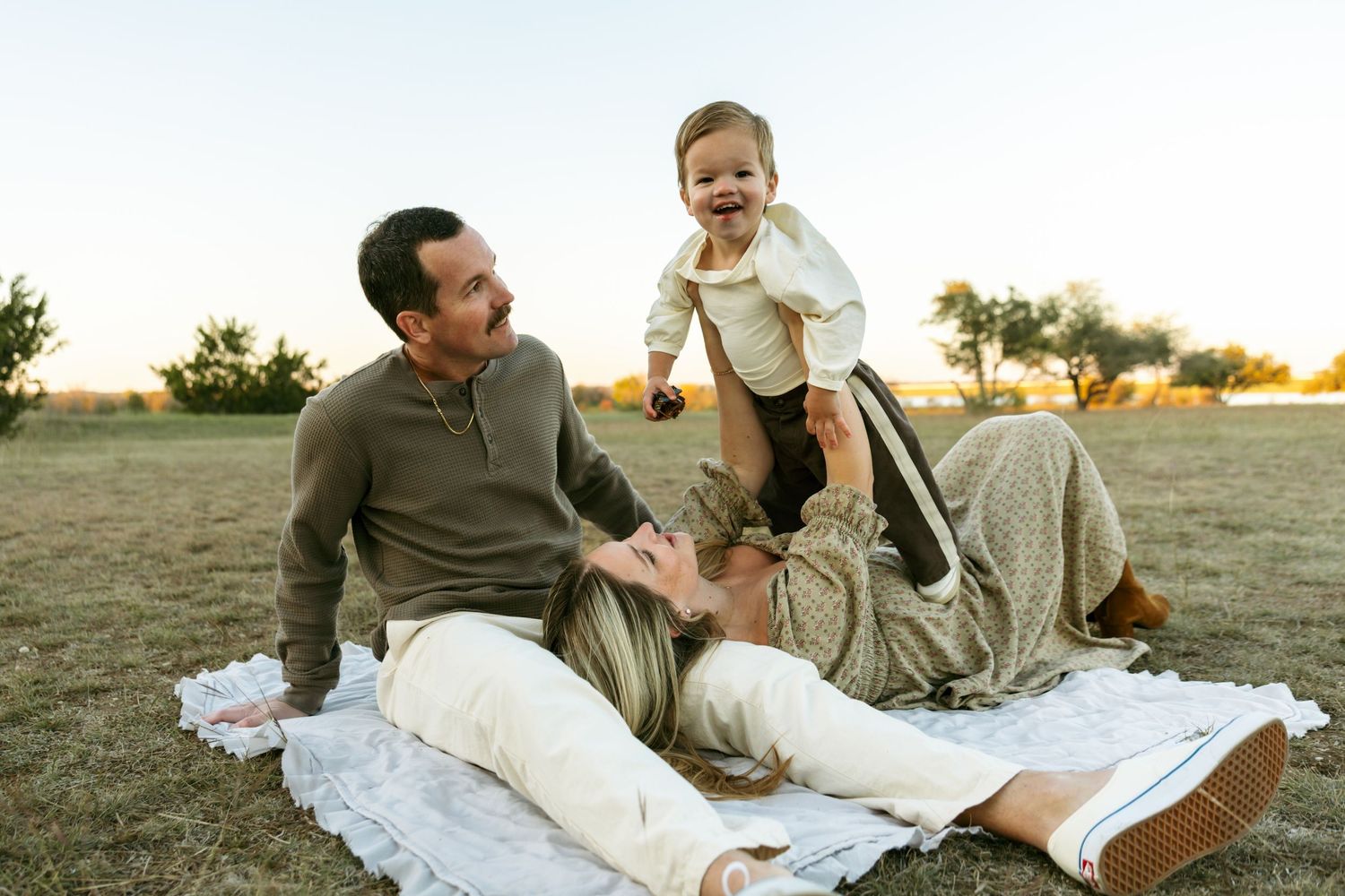 Happy family enjoys a golden hour picnic on a blanket outdoors, with parents playing with their laughing toddler.