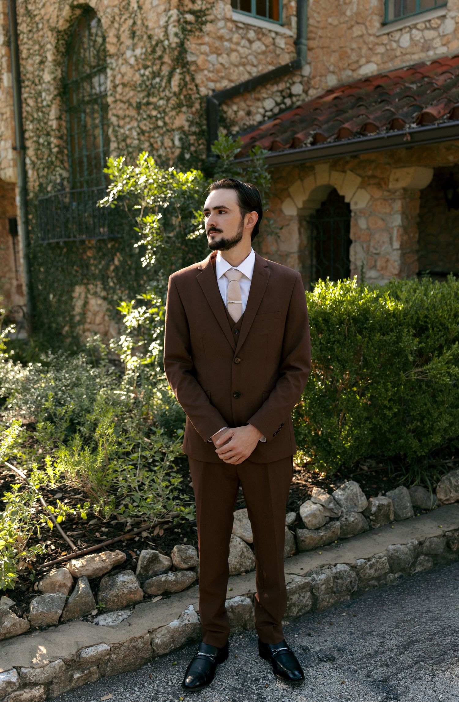 Dapper groom in brown three-piece suit standing before stone building surrounded by lush greenery.