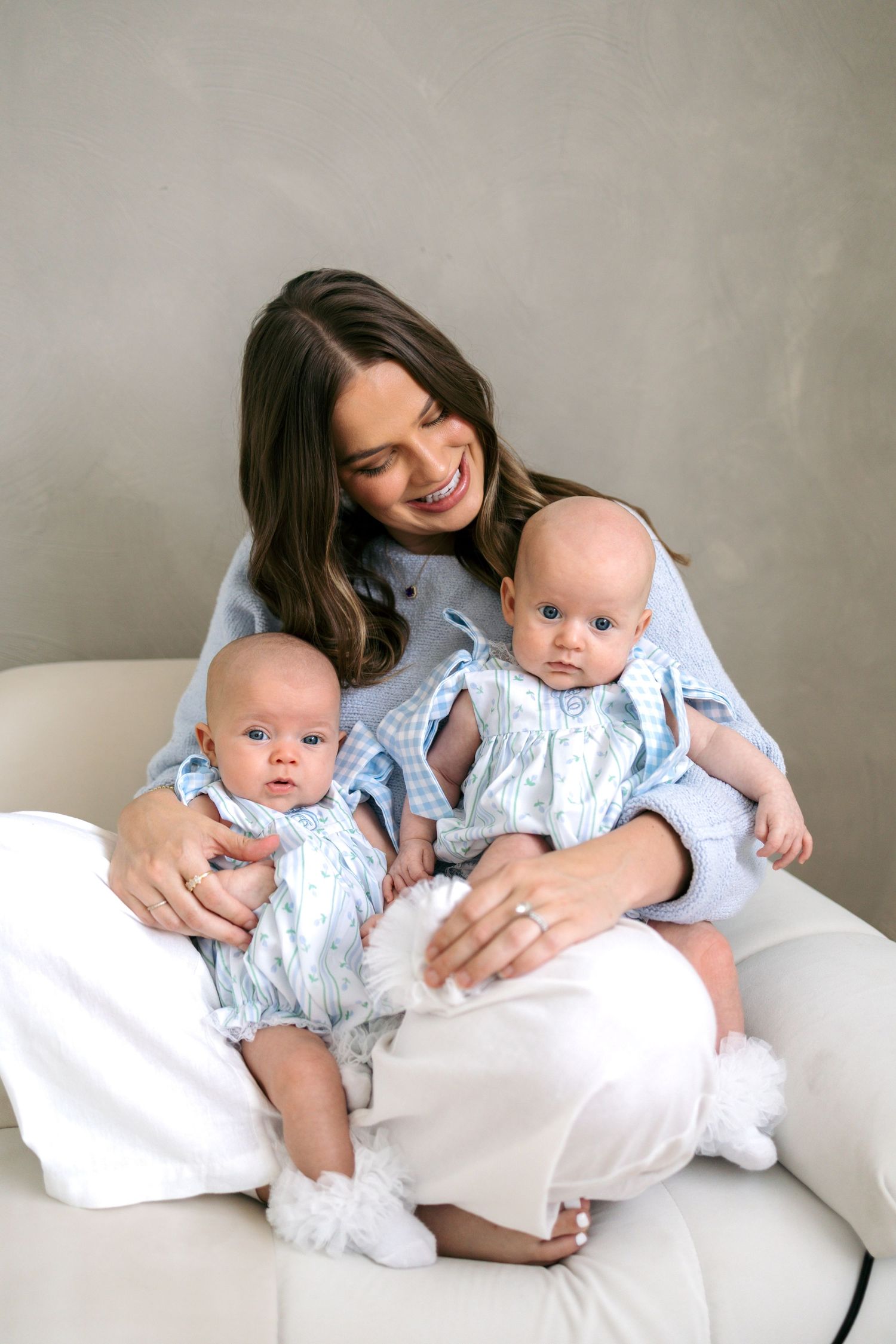 A smiling brunette woman holds two babies dressed in matching blue outfits on a white chair.