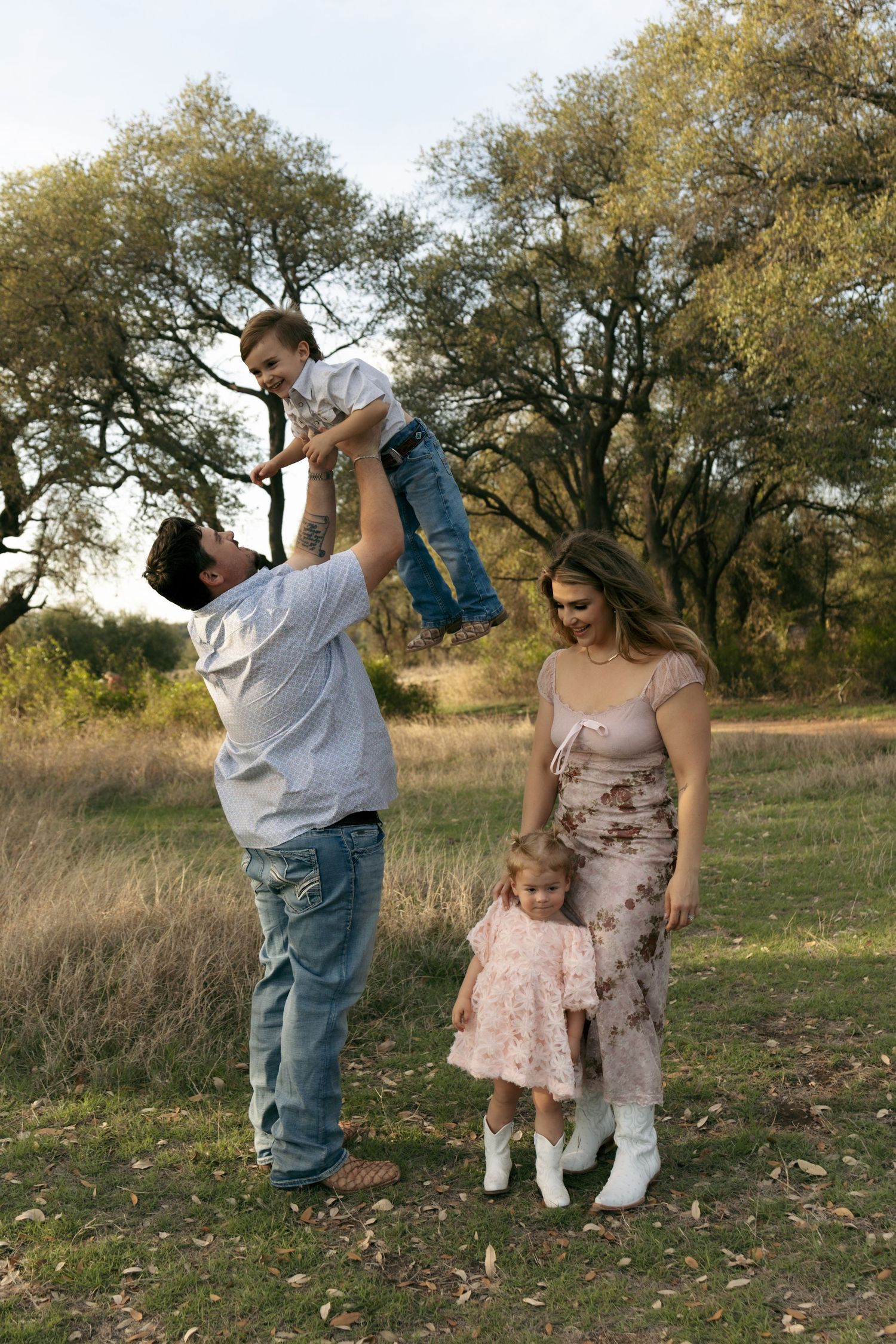 Father lifting baby boy in air while mother and daughter stand smiling in grassy field.