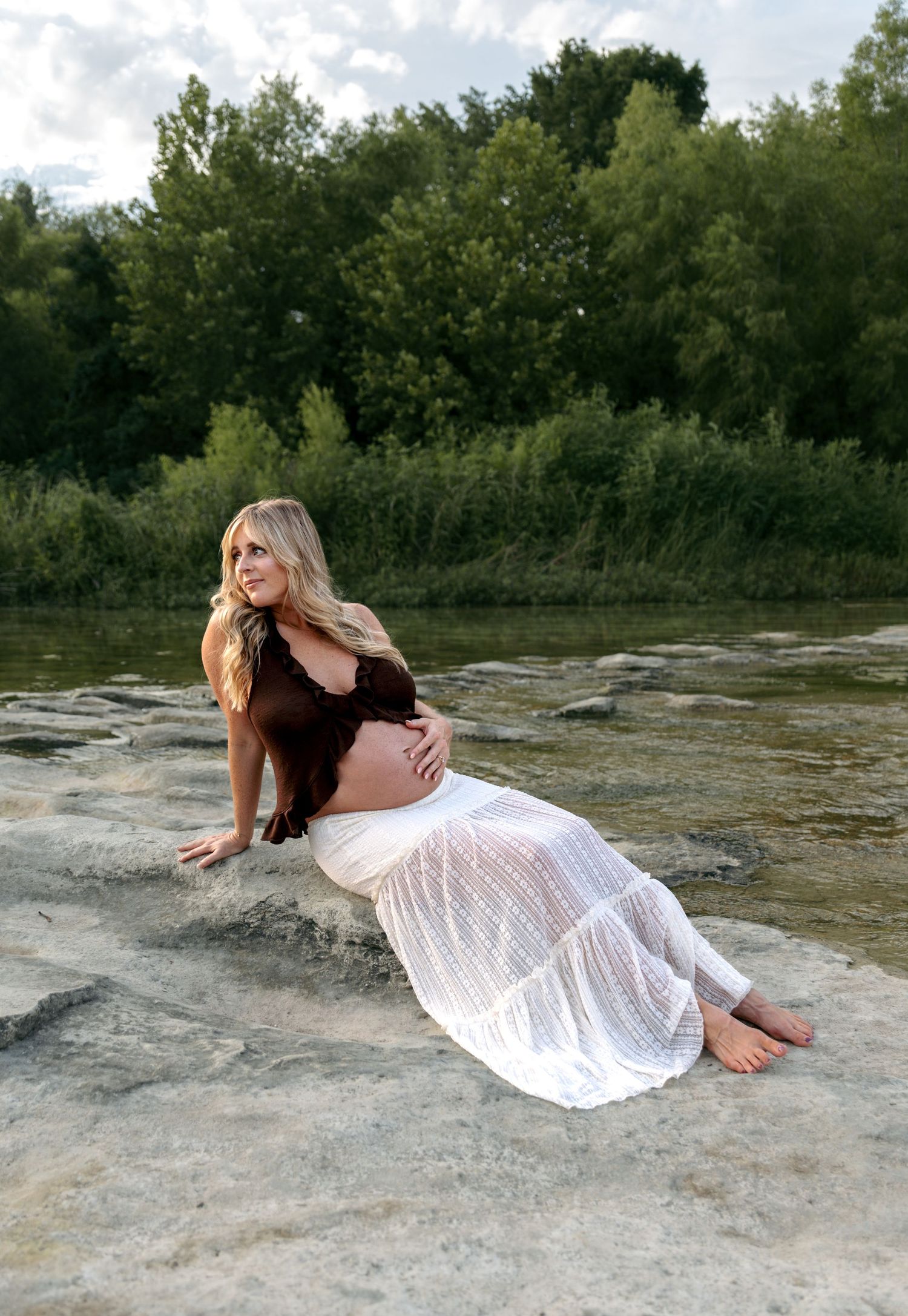 Pregnant woman in brown top and white skirt sitting on rocks near a river.