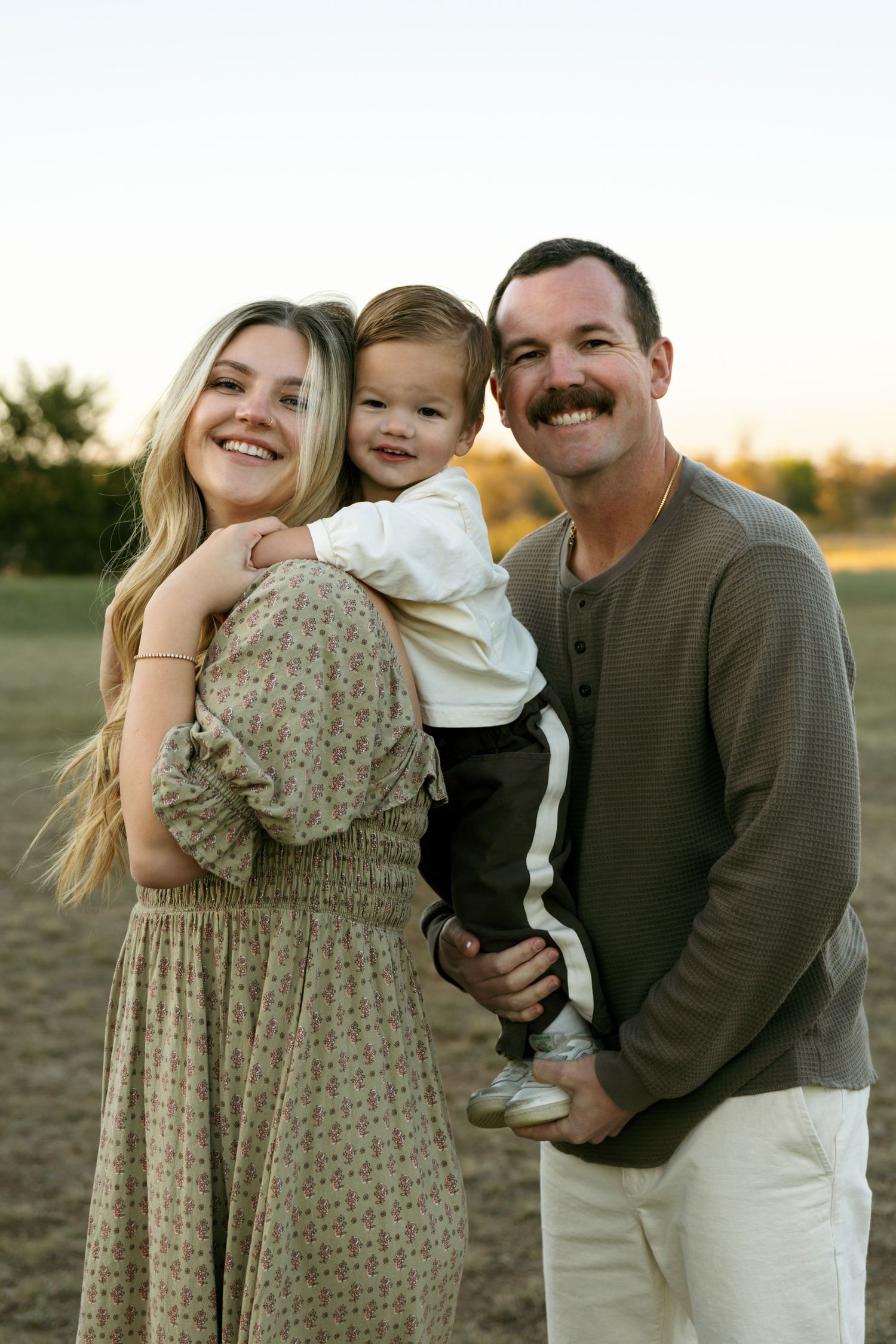 Smiling parents holding young toddler boy during golden hour outdoor family portrait session.