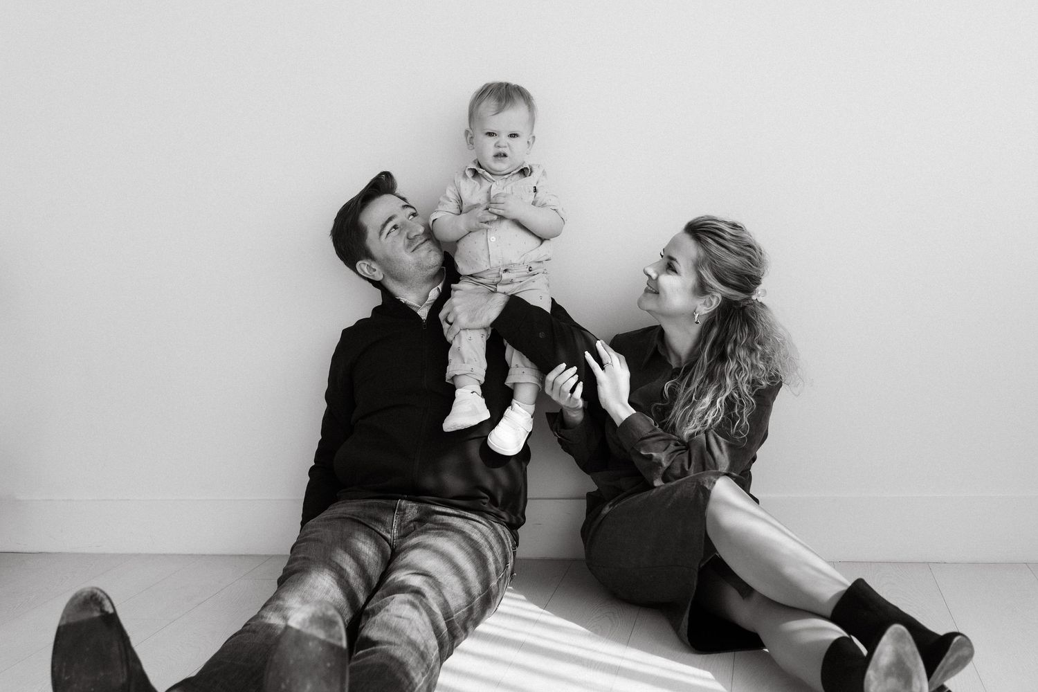 Black and white photo of parents sitting on floor laughing while holding happy baby between them.