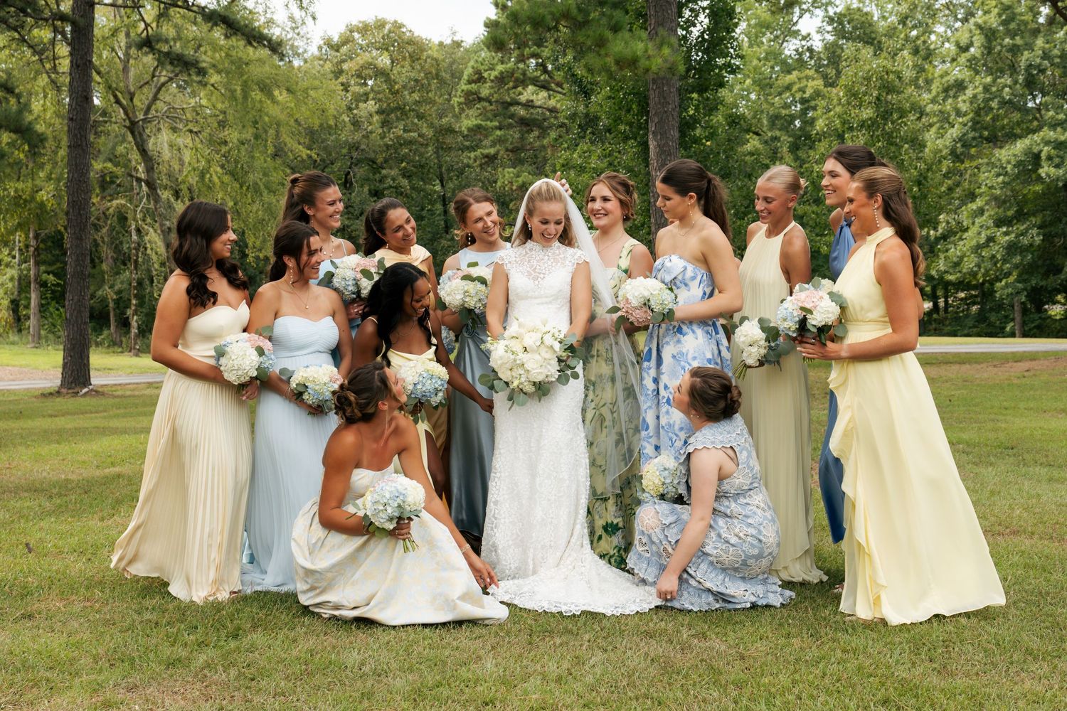 Bride surrounded by bridesmaids in mismatched pastel gowns holding white bouquets on a green lawn.