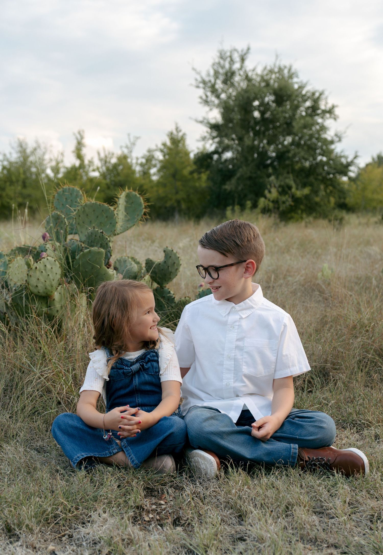 Two siblings, a boy in glasses and a girl in denim overalls, sit together smiling in a field near a cactus.
