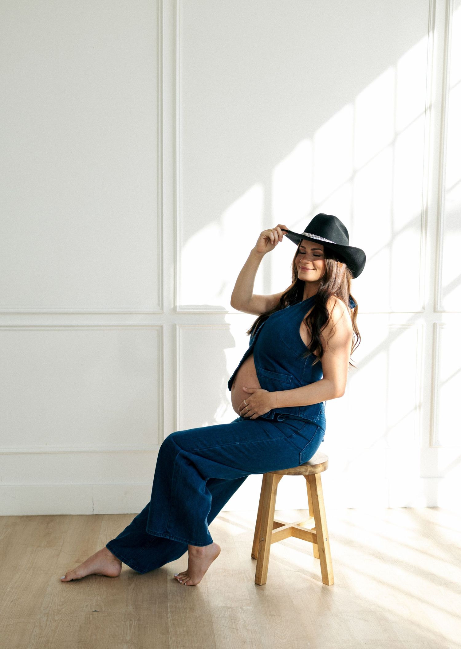 Pregnant woman in blue jumpsuit and black cowboy hat sitting on a wooden stool smiling.