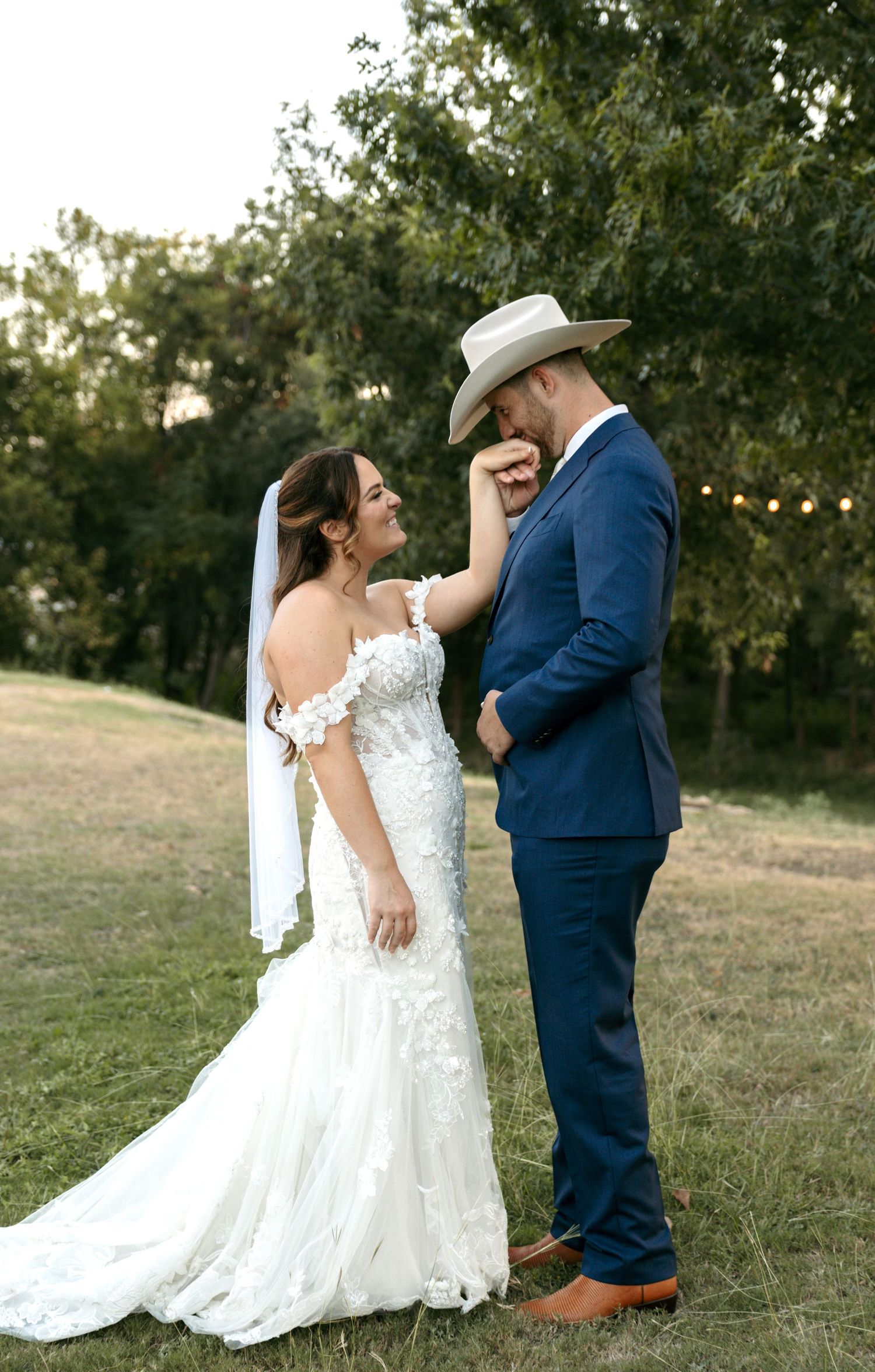 Bride in floral off-shoulder gown adjusting grooms tan cowboy hat in an outdoor field.