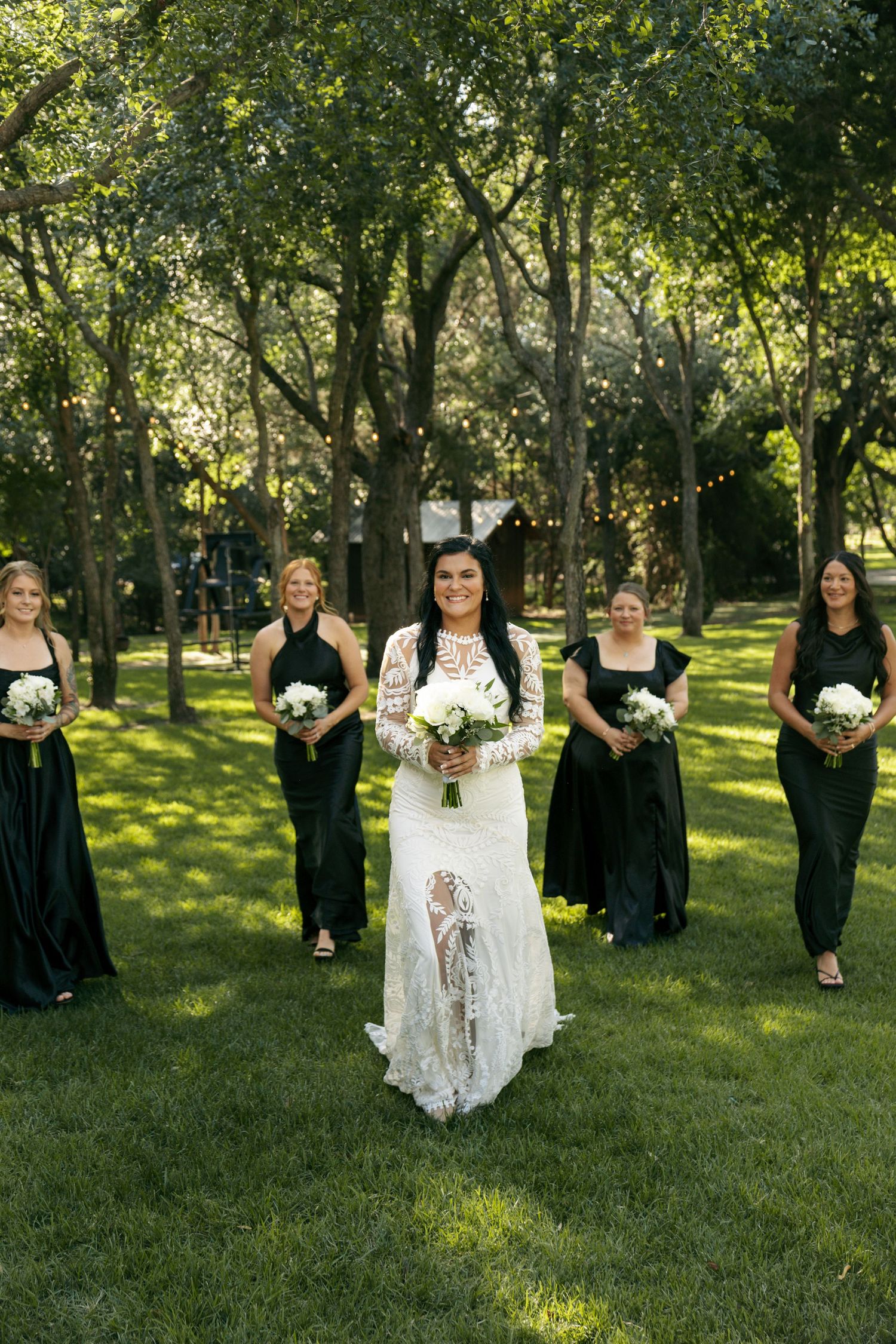 A bride in lace leads her bridesmaids in black dresses across a green lawn surrounded by string-lit trees.