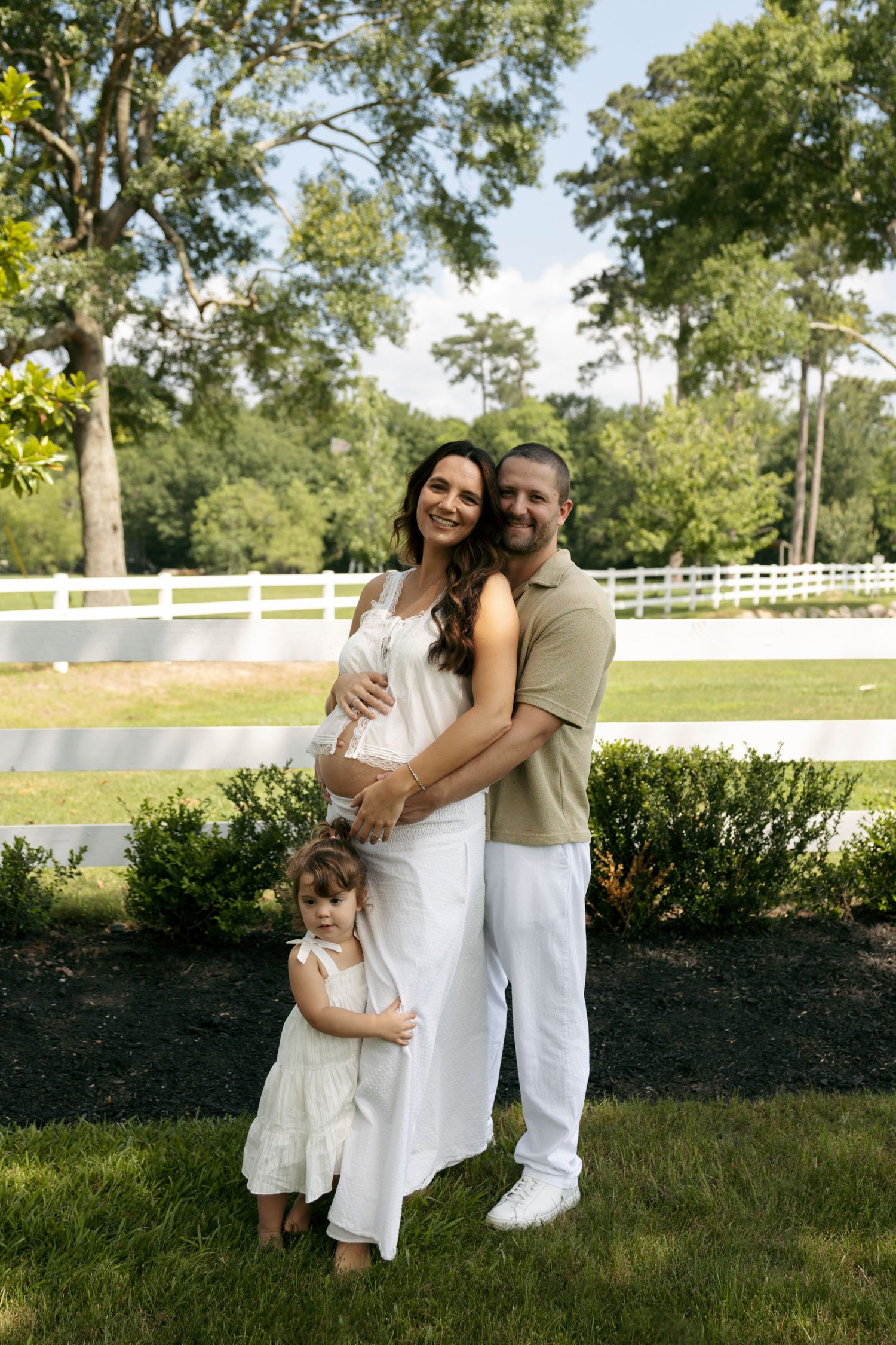 A pregnant woman in white stands with her husband and young daughter in a sunny garden with a white fence.