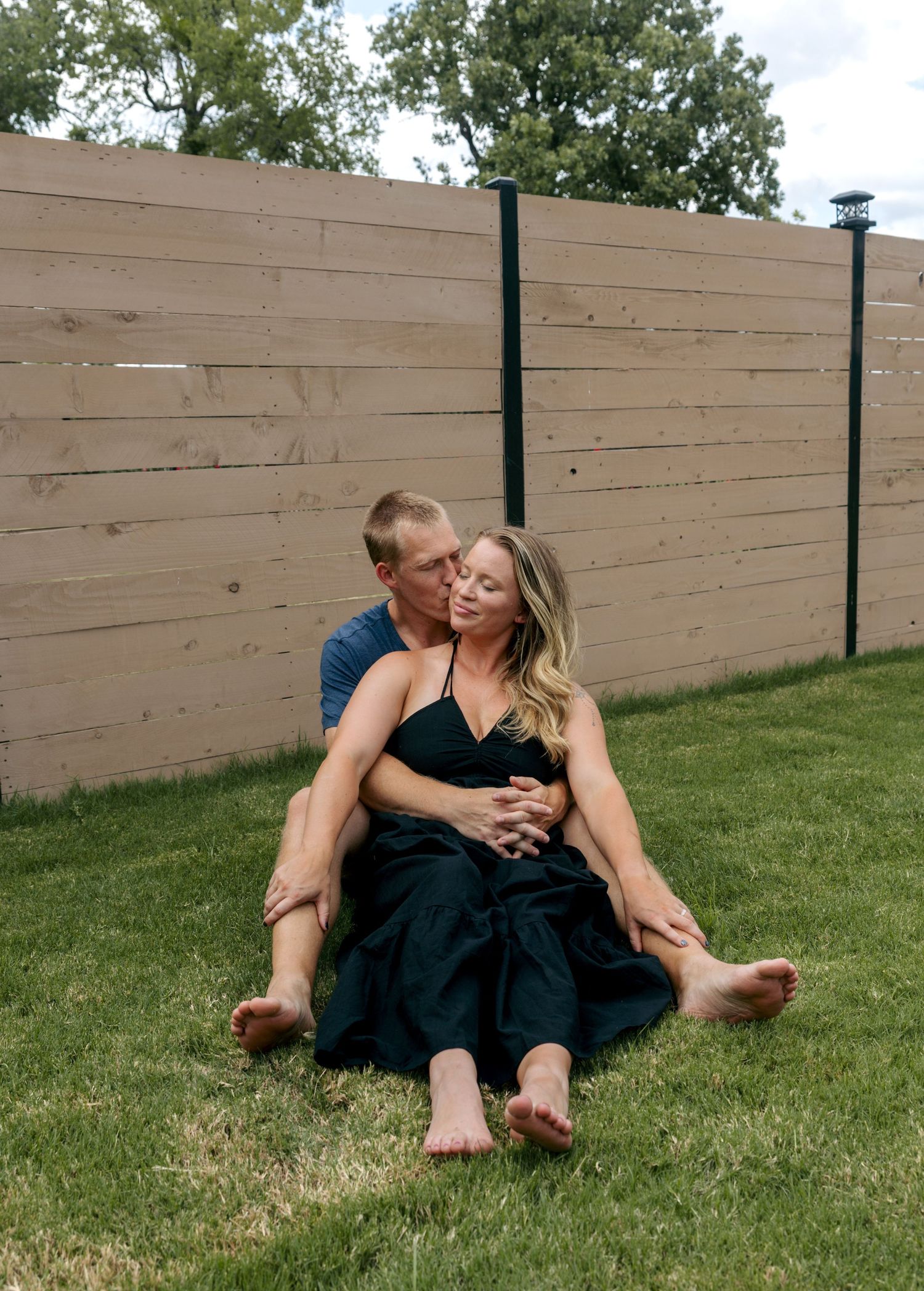 Couple sitting together on grass in backyard, sharing a romantic kiss.