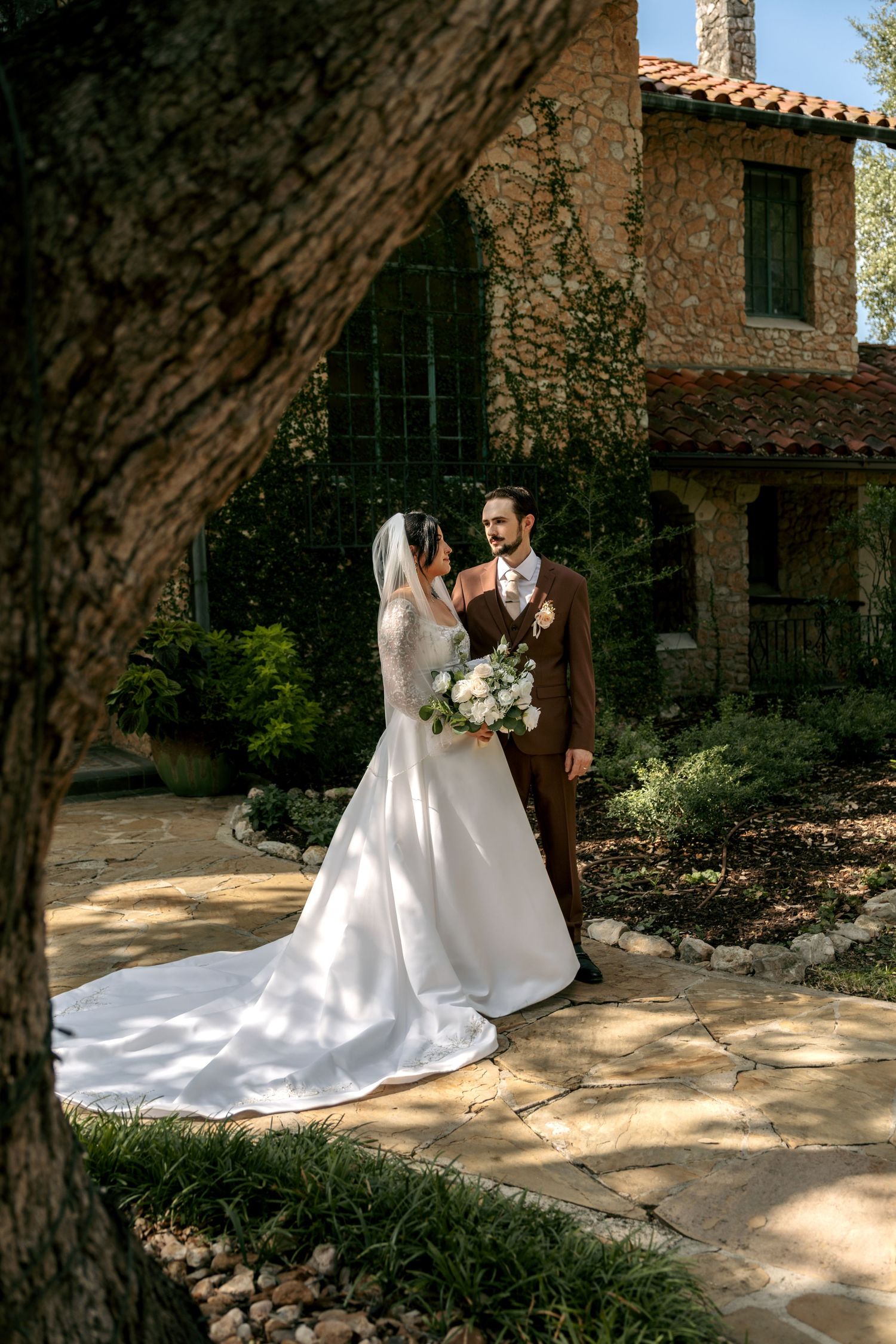 Bride in cathedral veil and groom in brown suit pose in garden of elegant stone estate.
