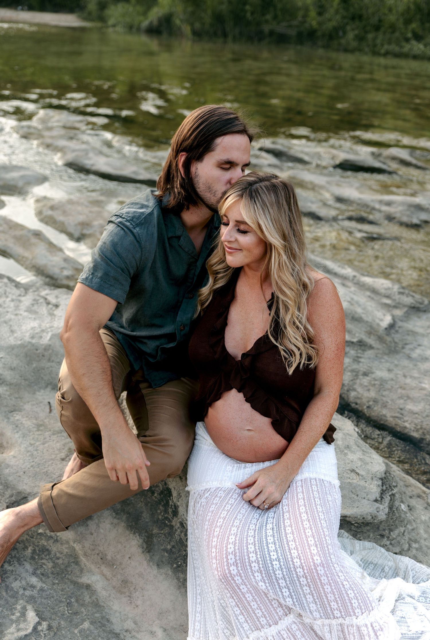 Couple sharing intimate moment on rocky riverbank, pregnant woman in white skirt and brown top.