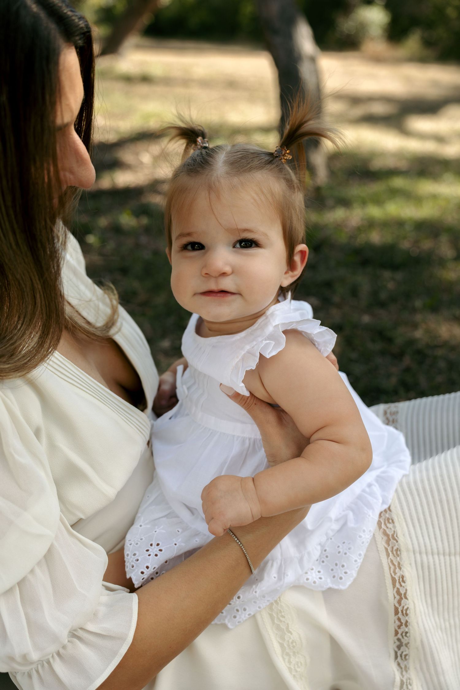 Mother in white dress holding adorable baby girl with pigtails in white eyelet dress outdoors.
