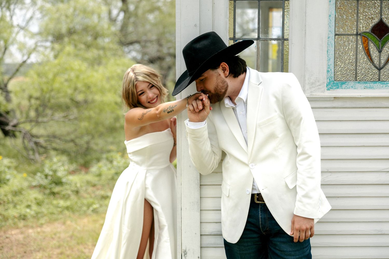Couple in western-style wedding attire sharing a romantic moment outside a rustic white building.