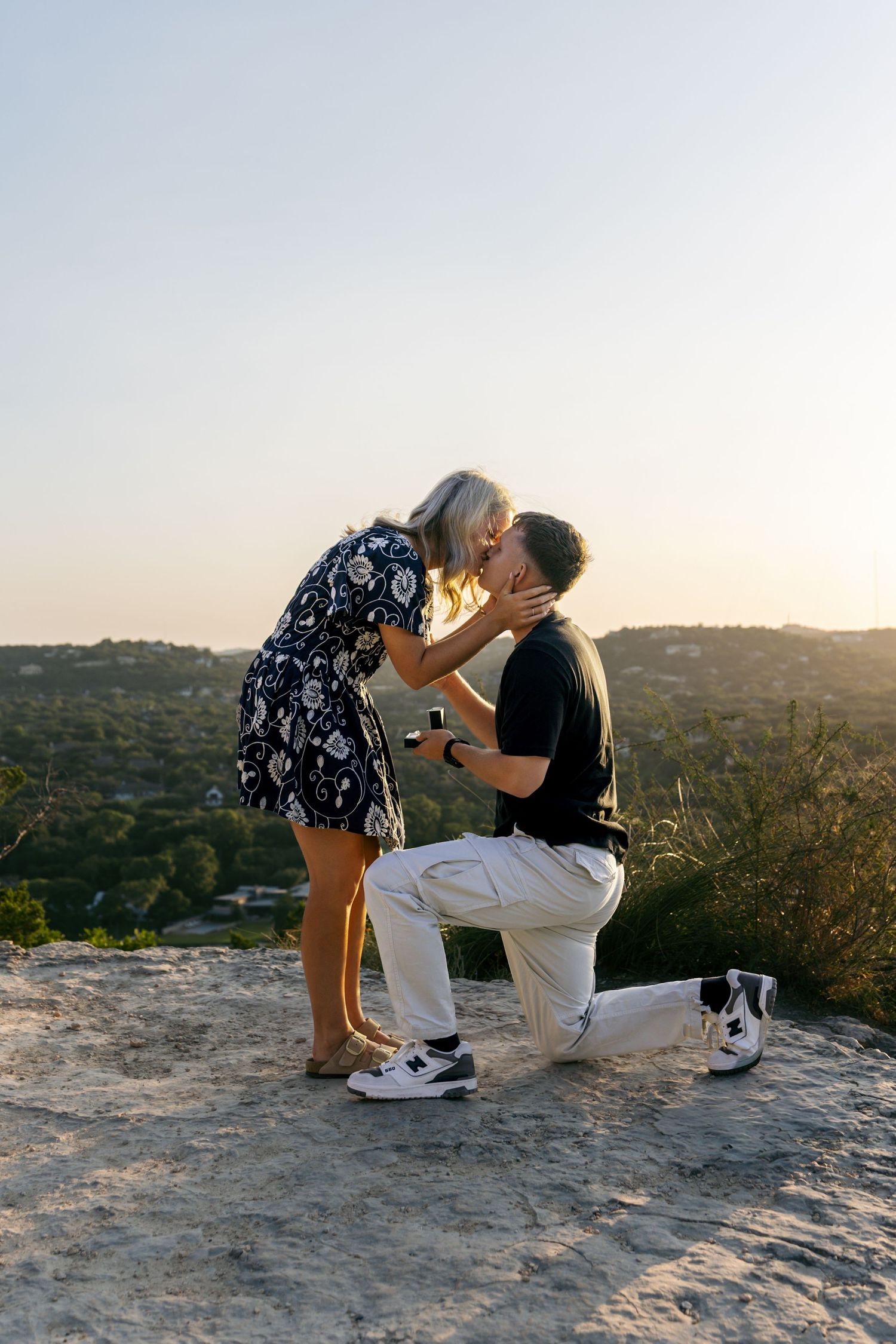 Man proposing on one knee to woman at golden hour on a rocky hilltop overlook.