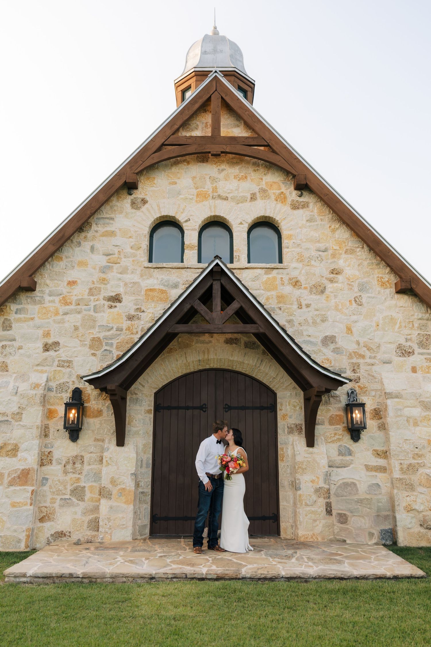 Newlywed couple kissing in front of a rustic stone chapel with wooden doors.