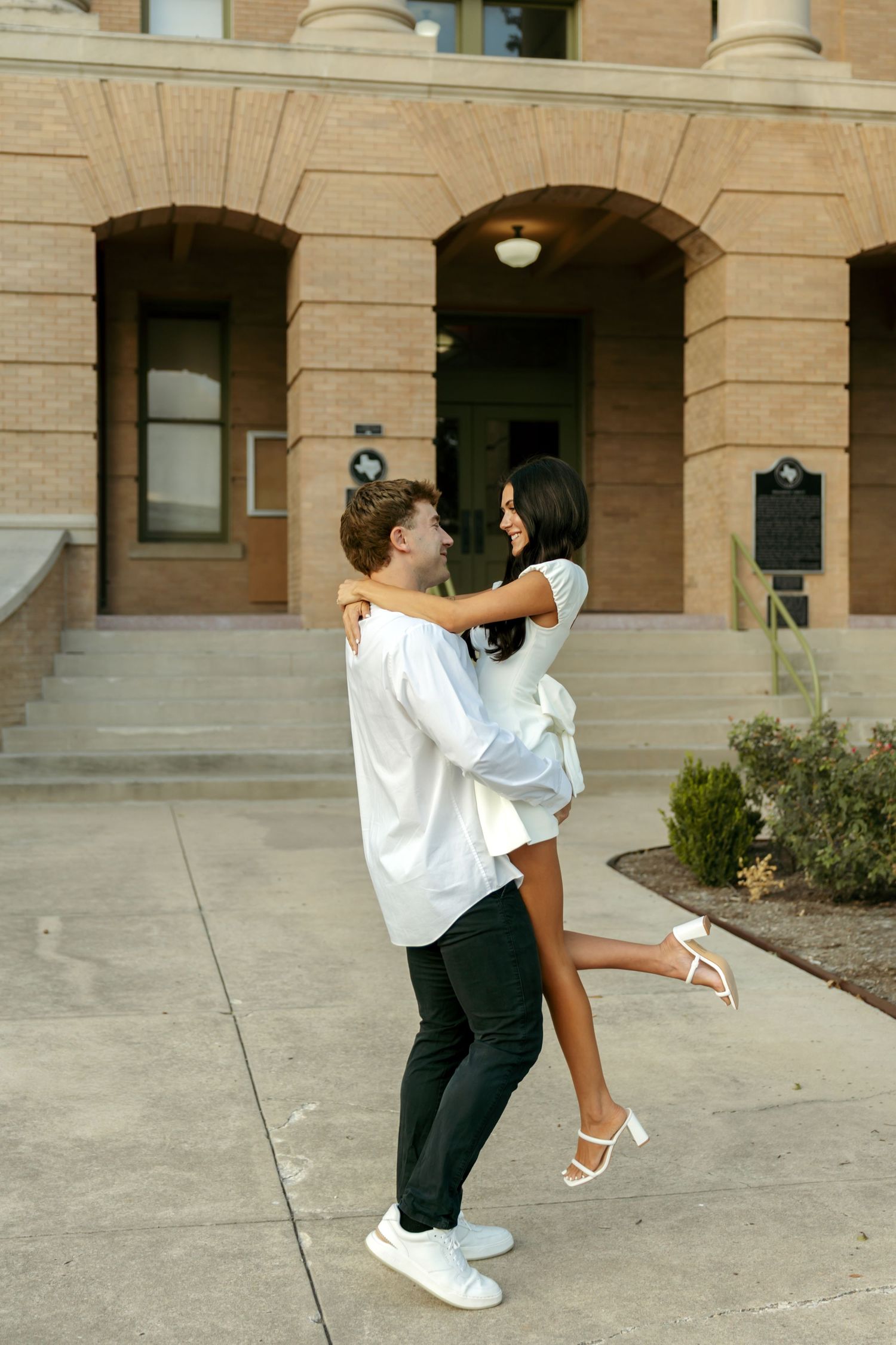Man lifting woman in white dress outside a brick building, both smiling joyfully.