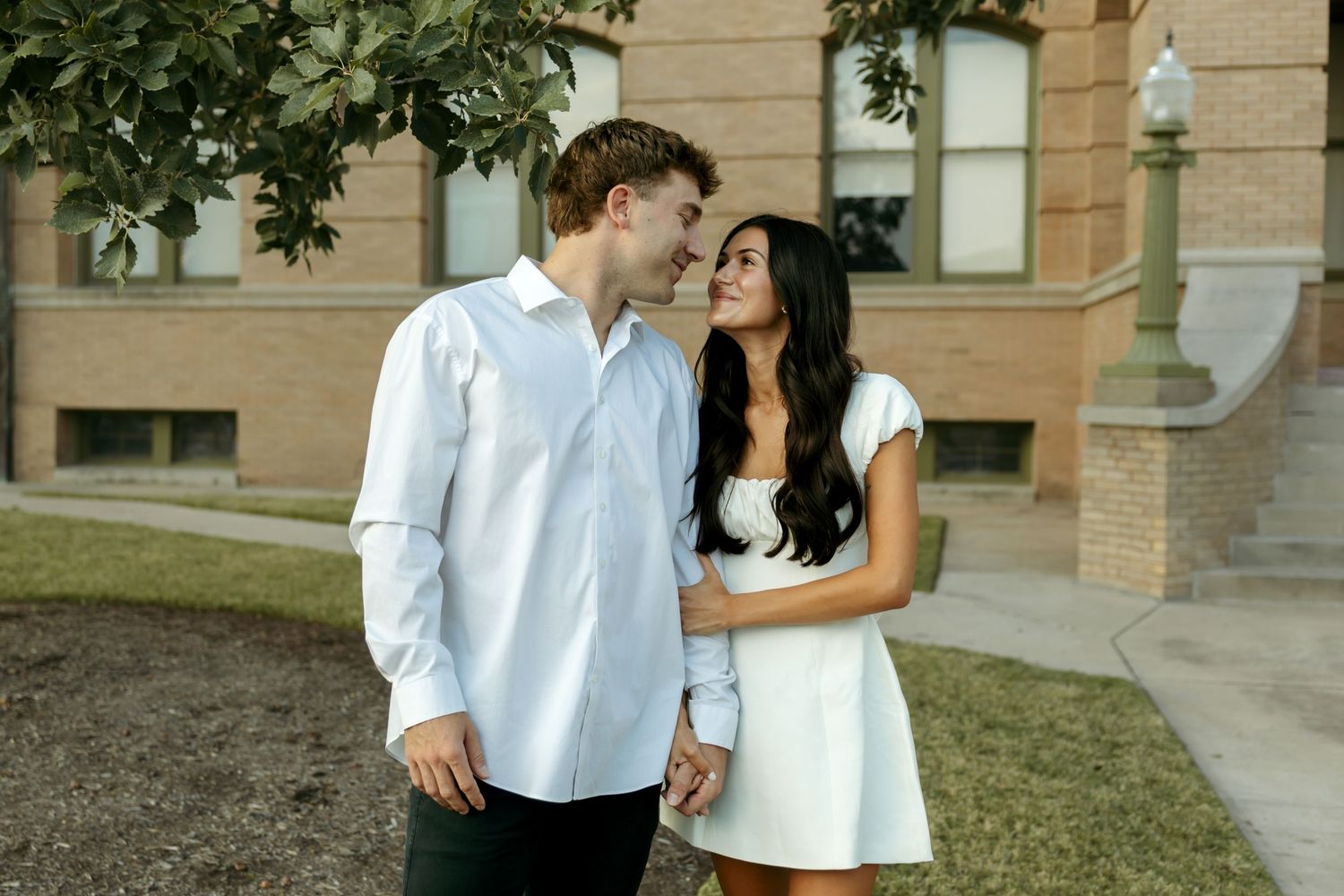 Happy couple shares a romantic moment outdoors near a brick building during their engagement photo session.
