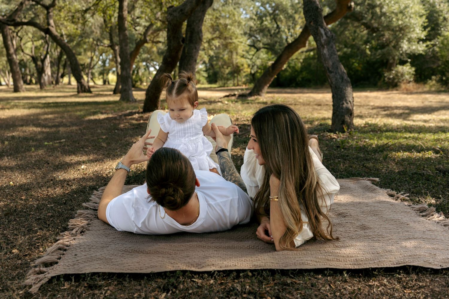 Young family relaxing on blanket in shaded park, parents playing with toddler in white dress.