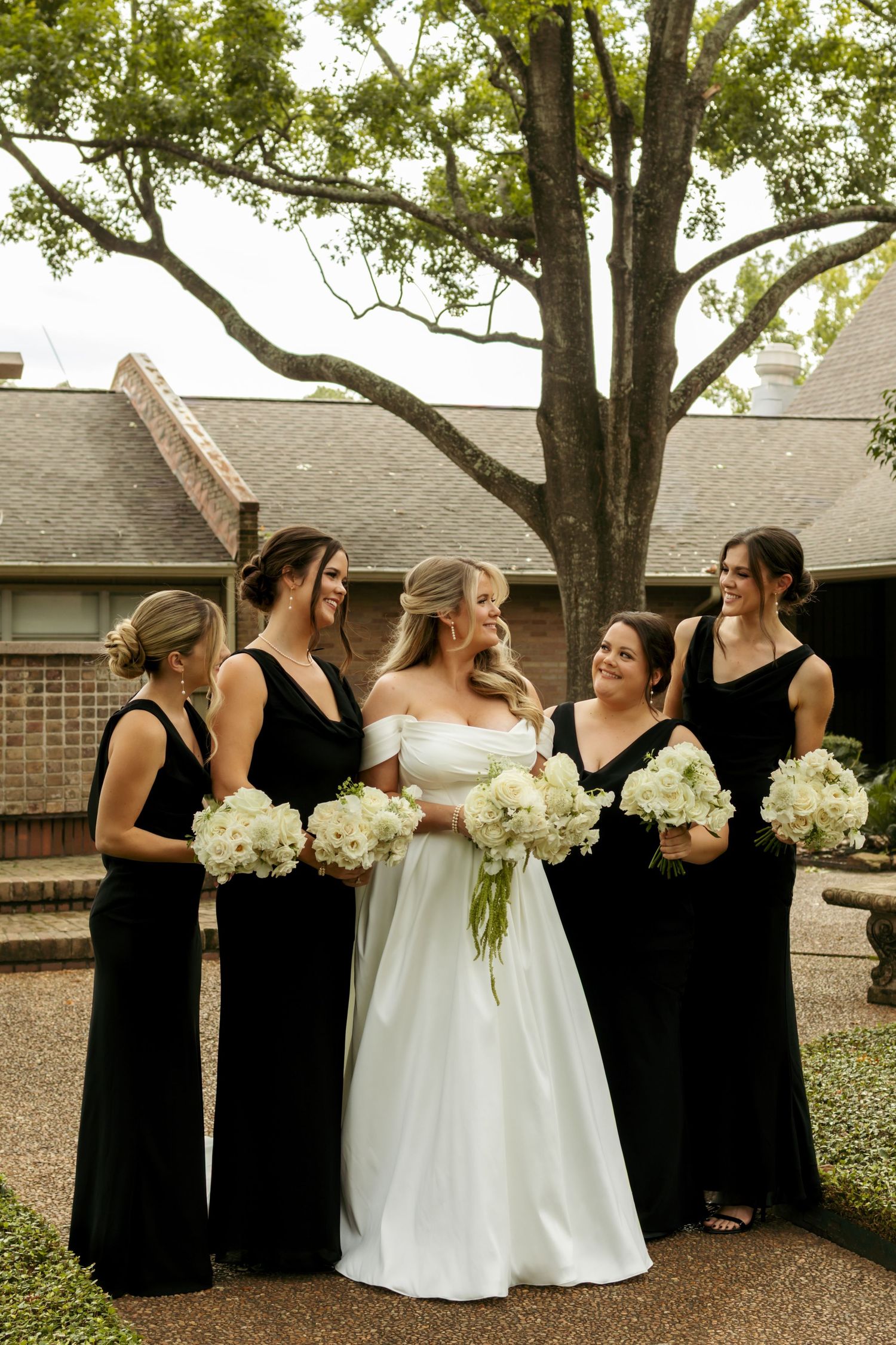 Bride in white off-shoulder gown stands with four bridesmaids in black dresses holding white floral bouquets.
