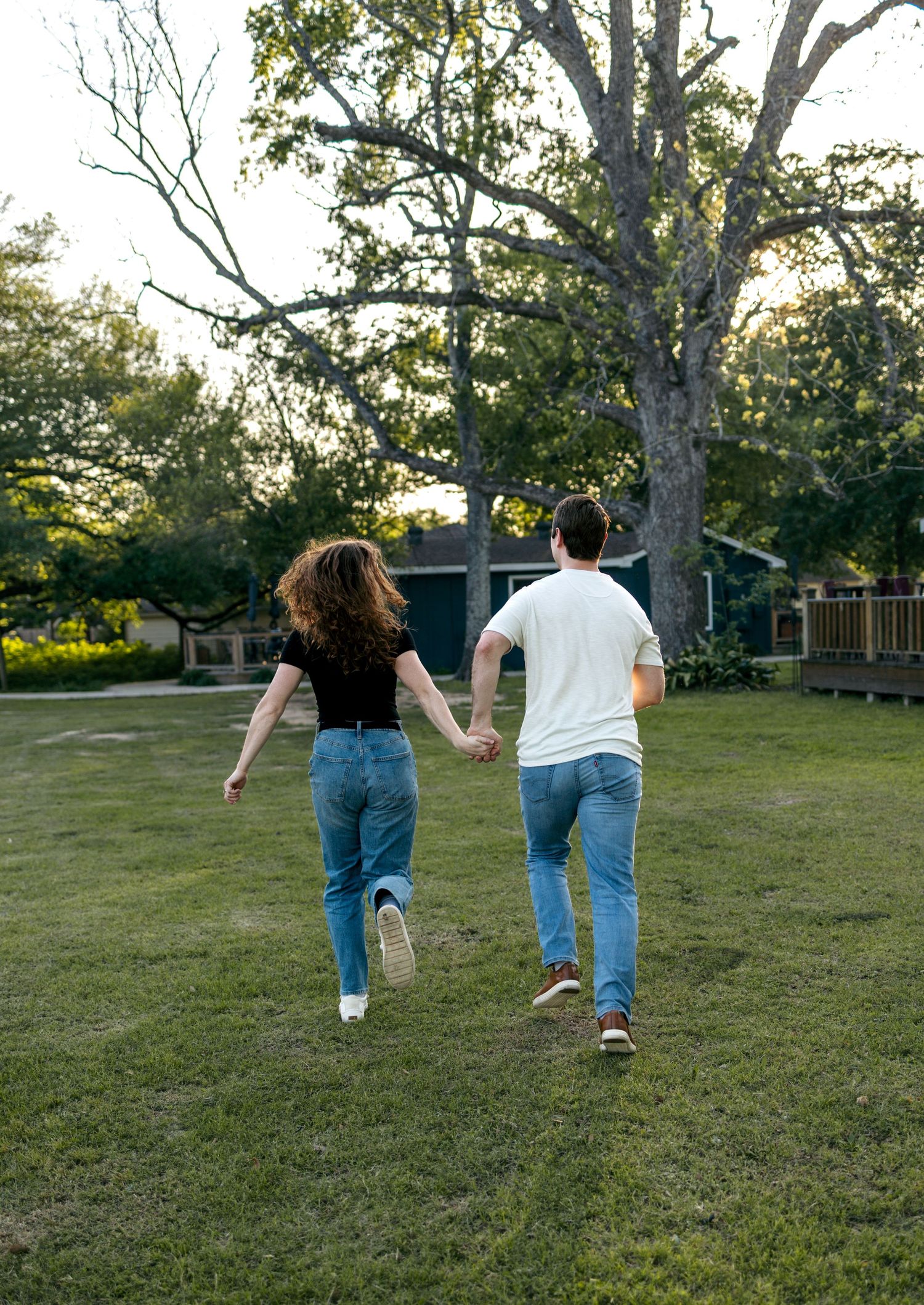 Couple holding hands running through green yard toward large tree.