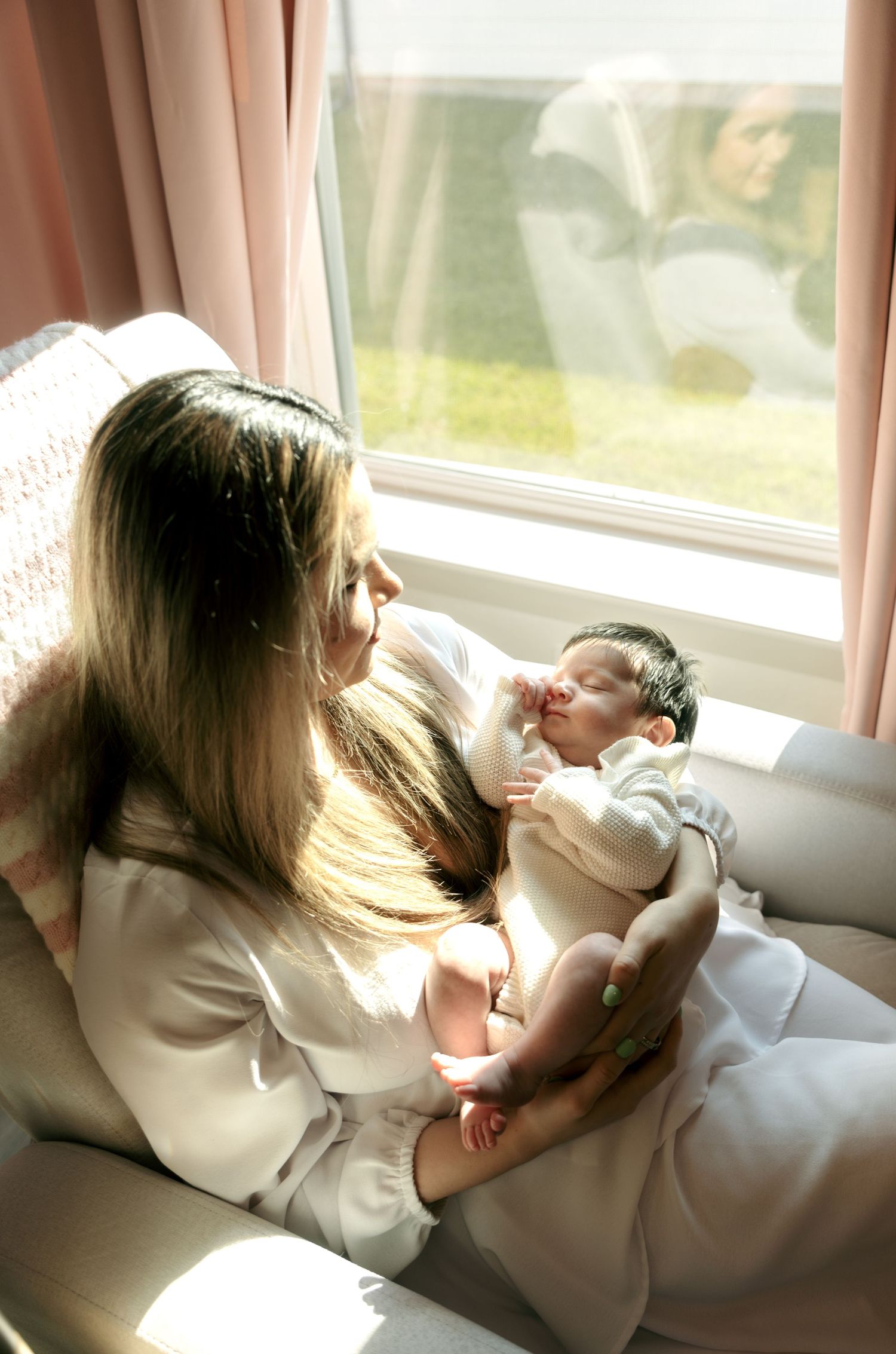 Mother gazes lovingly at her newborn baby while sitting in a sunlit chair by a window.