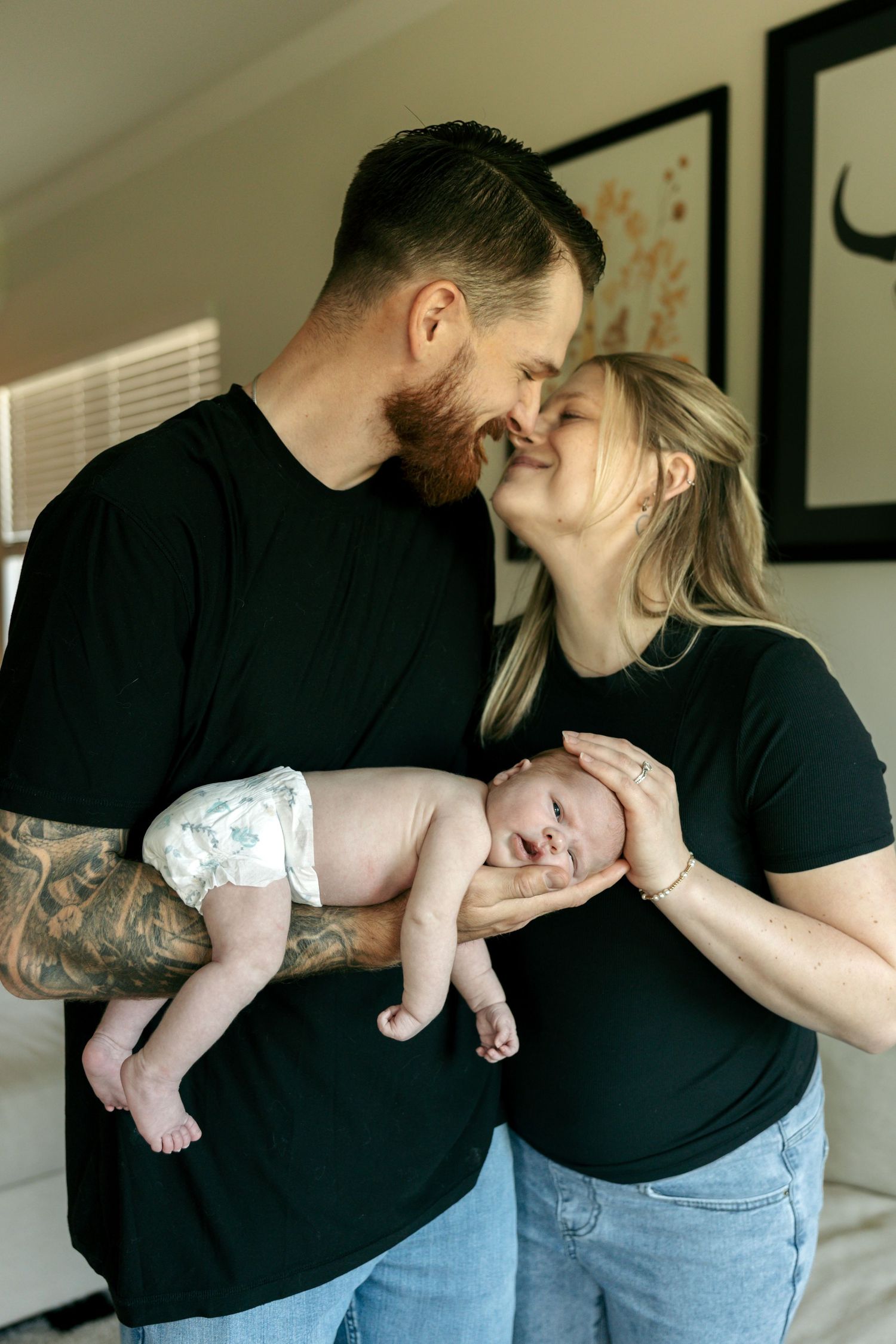 Couple in matching black shirts sharing a kiss while holding their newborn baby.
