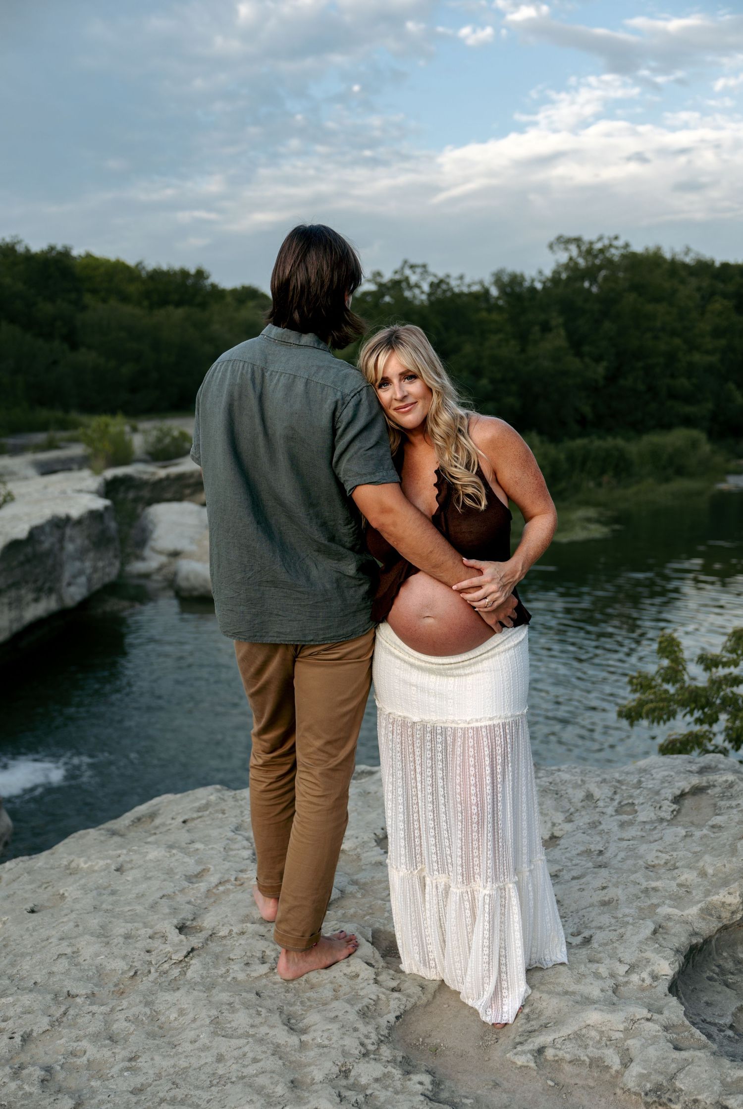 Pregnant woman in white skirt smiles back at camera while partner embraces her by a river.