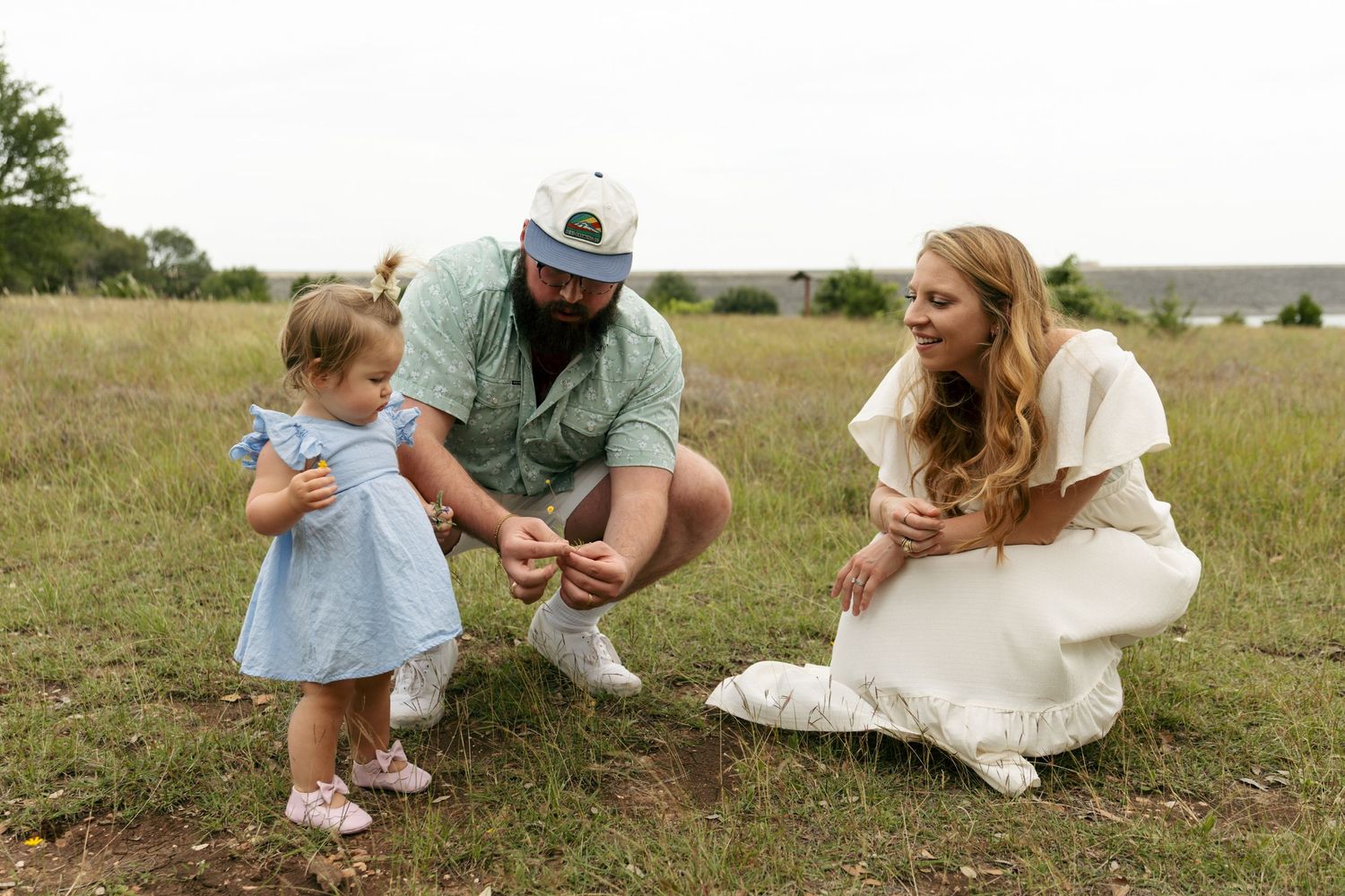 Young family with toddler girl in blue dress exploring grassy field together outdoors.