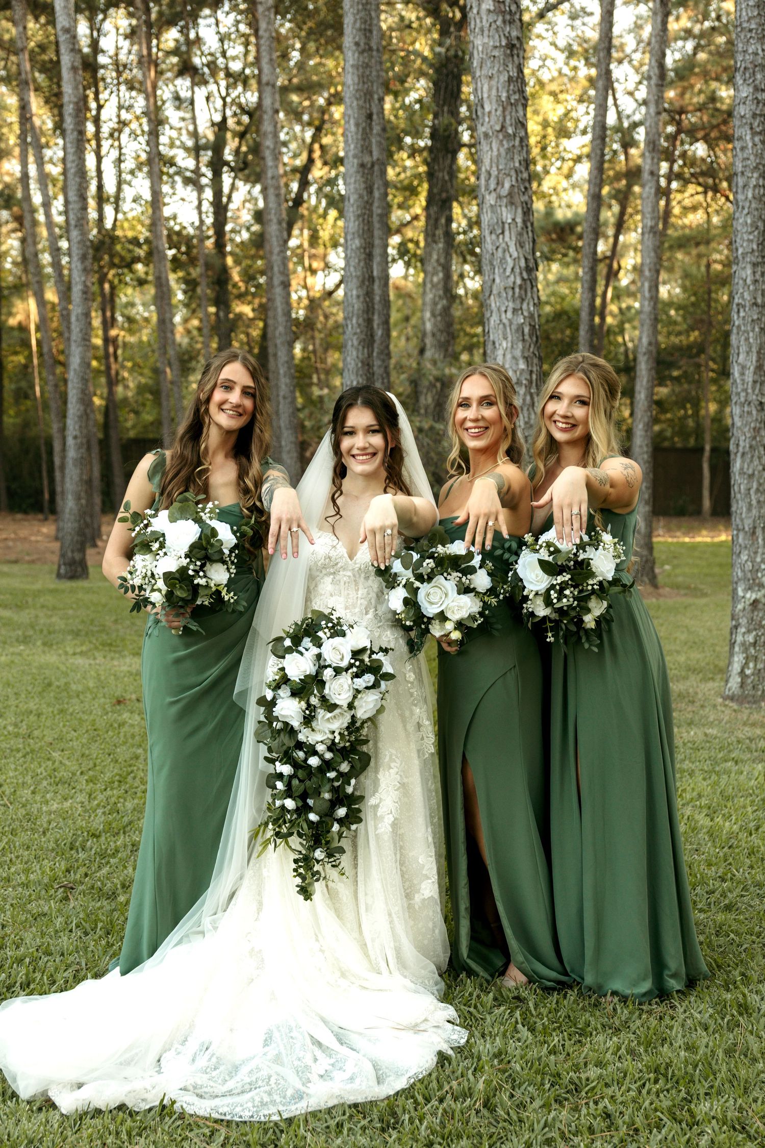 Bride in white lace gown with three bridesmaids in green dresses showing off rings outdoors.