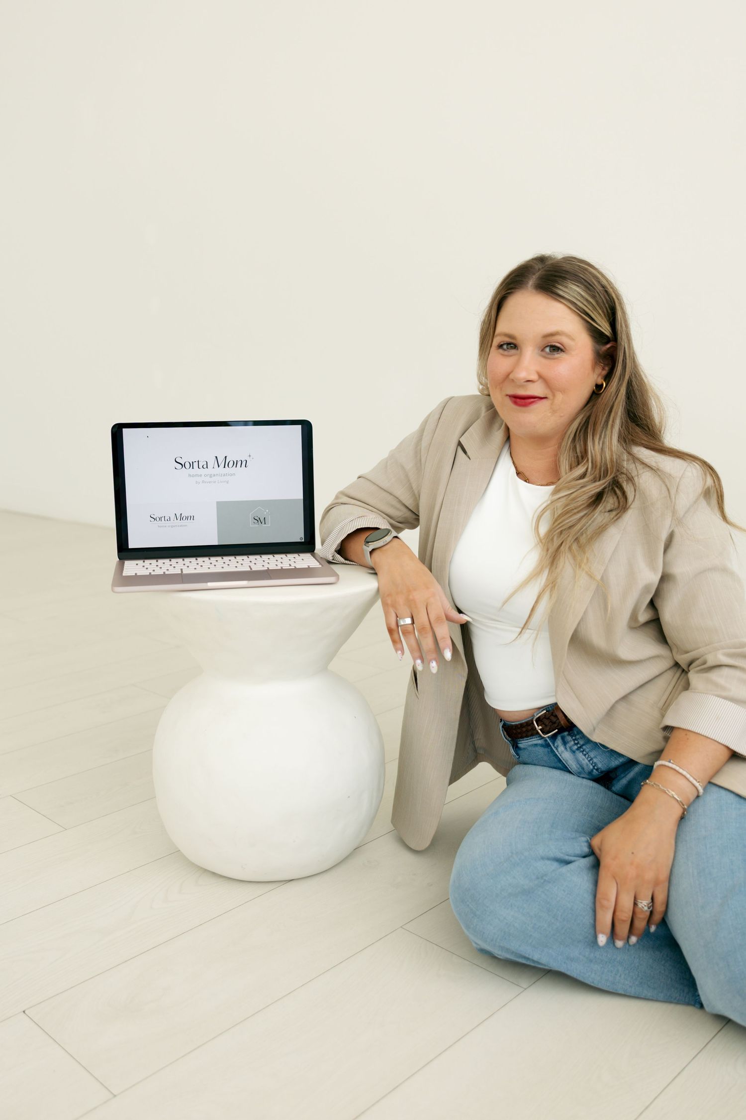 Smiling blonde businesswoman in beige blazer posing with laptop displaying Sorta Mom website on white floor.