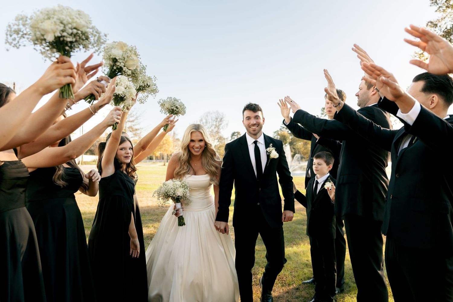 Wedding party celebrates with raised hands as bride and groom walk through an arch of cheering guests outdoors.