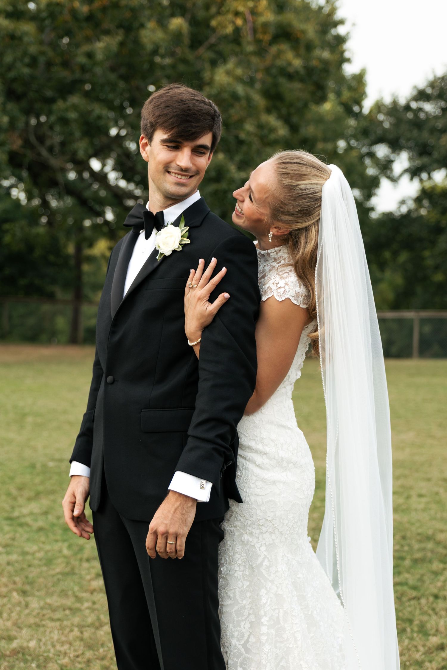 Bride in lace gown and veil embraces groom in black tuxedo outdoors on their wedding day.