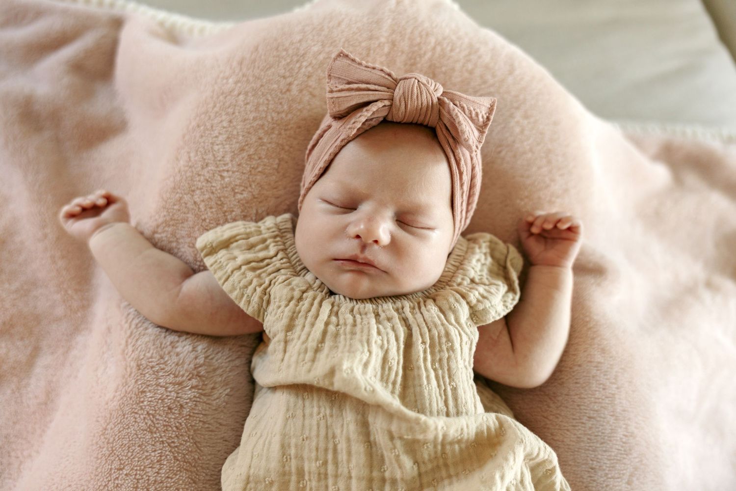 Sleeping newborn baby girl in yellow dress with pink bow headband on pink blanket.
