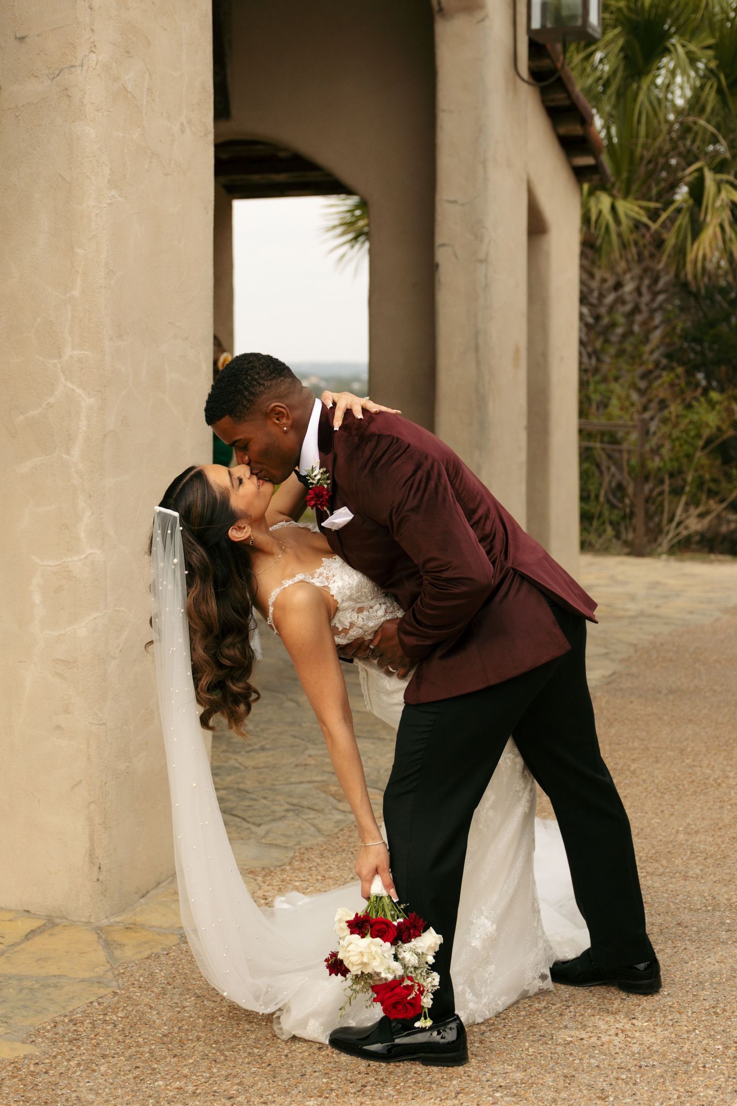 Bride and groom share a romantic dip kiss outdoors at their wedding venue with lush greenery in the background.