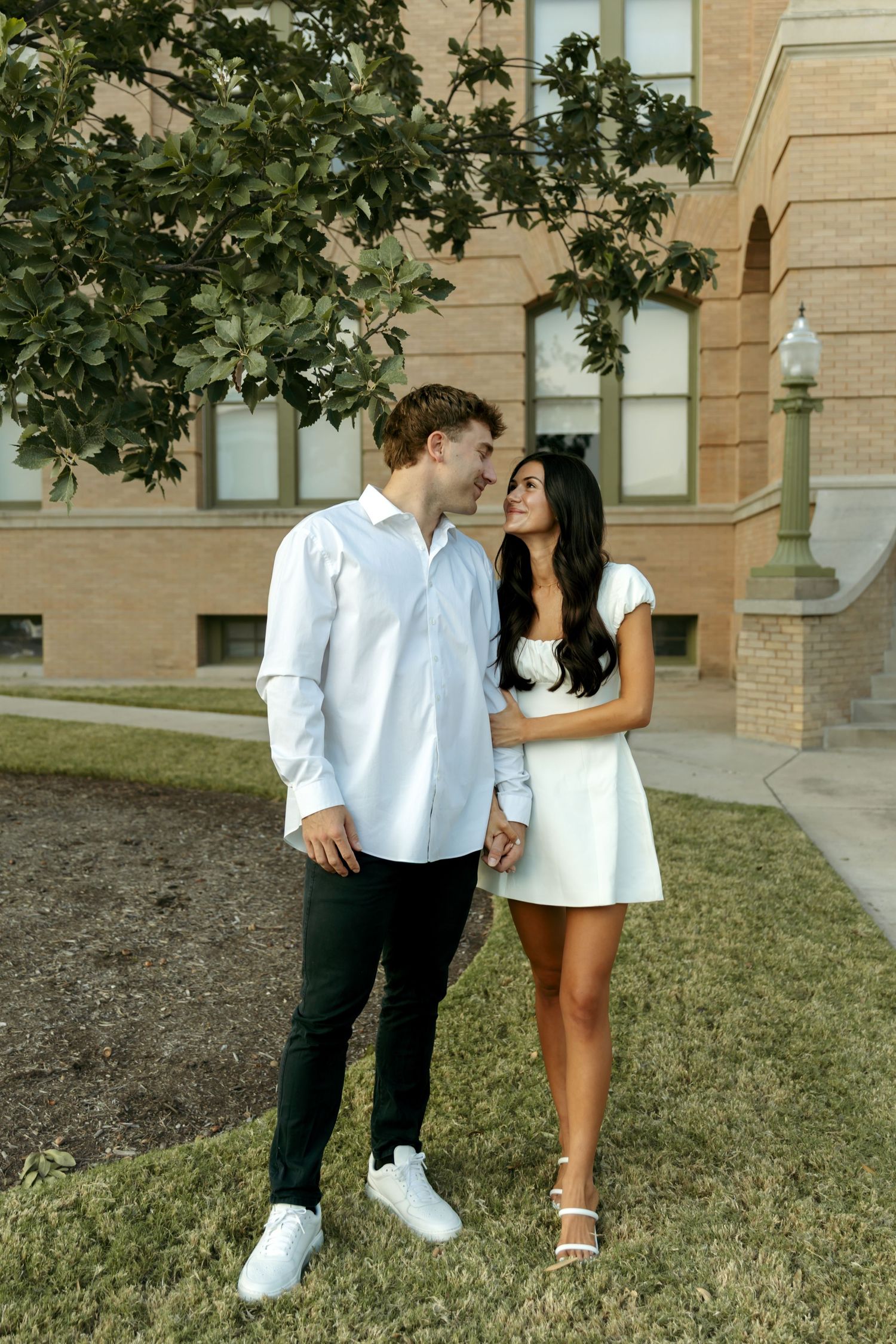 Young couple holding hands and smiling under a tree near a brick building.