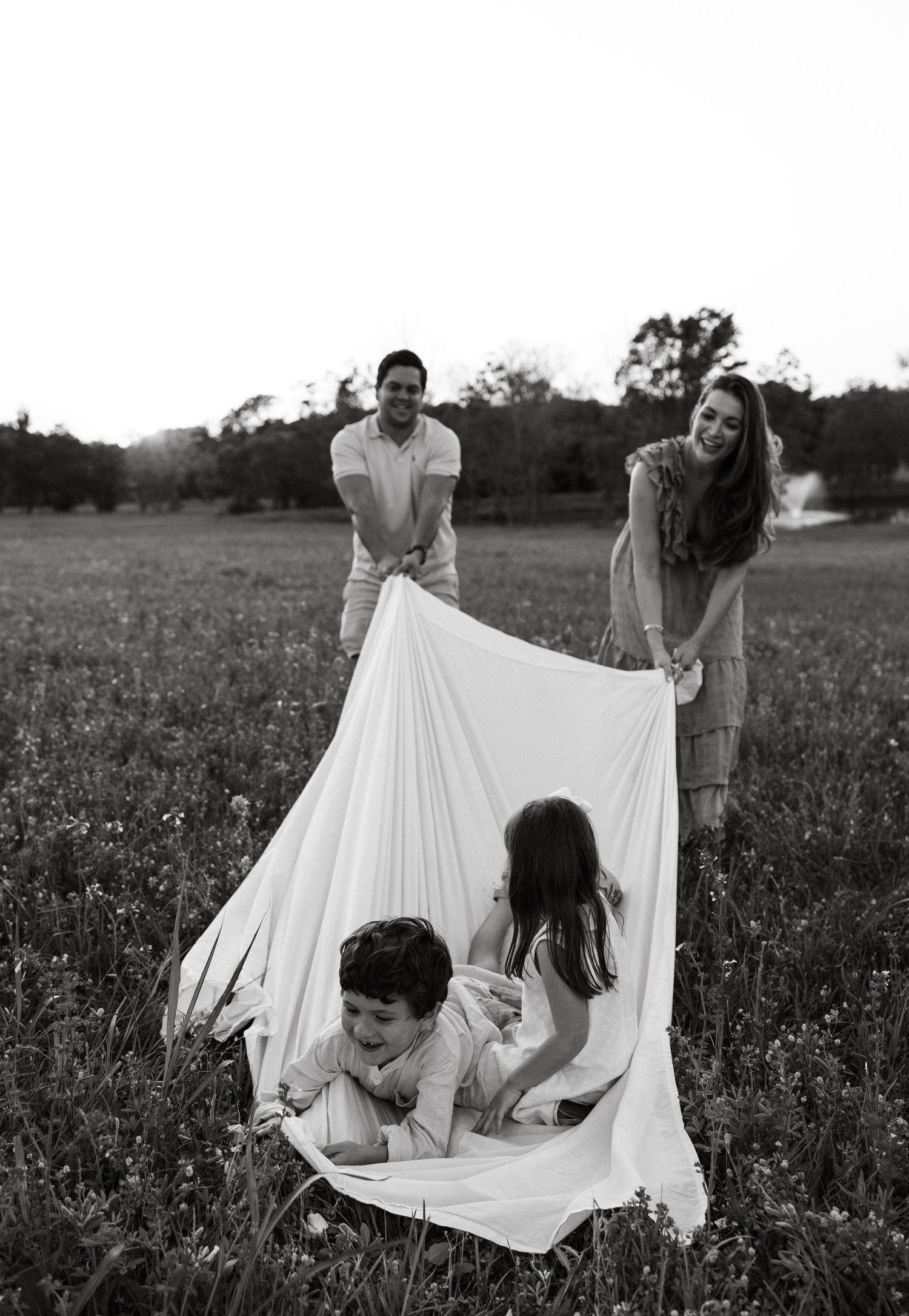 Black and white photo of parents swinging children in a white sheet in an open grassy field.