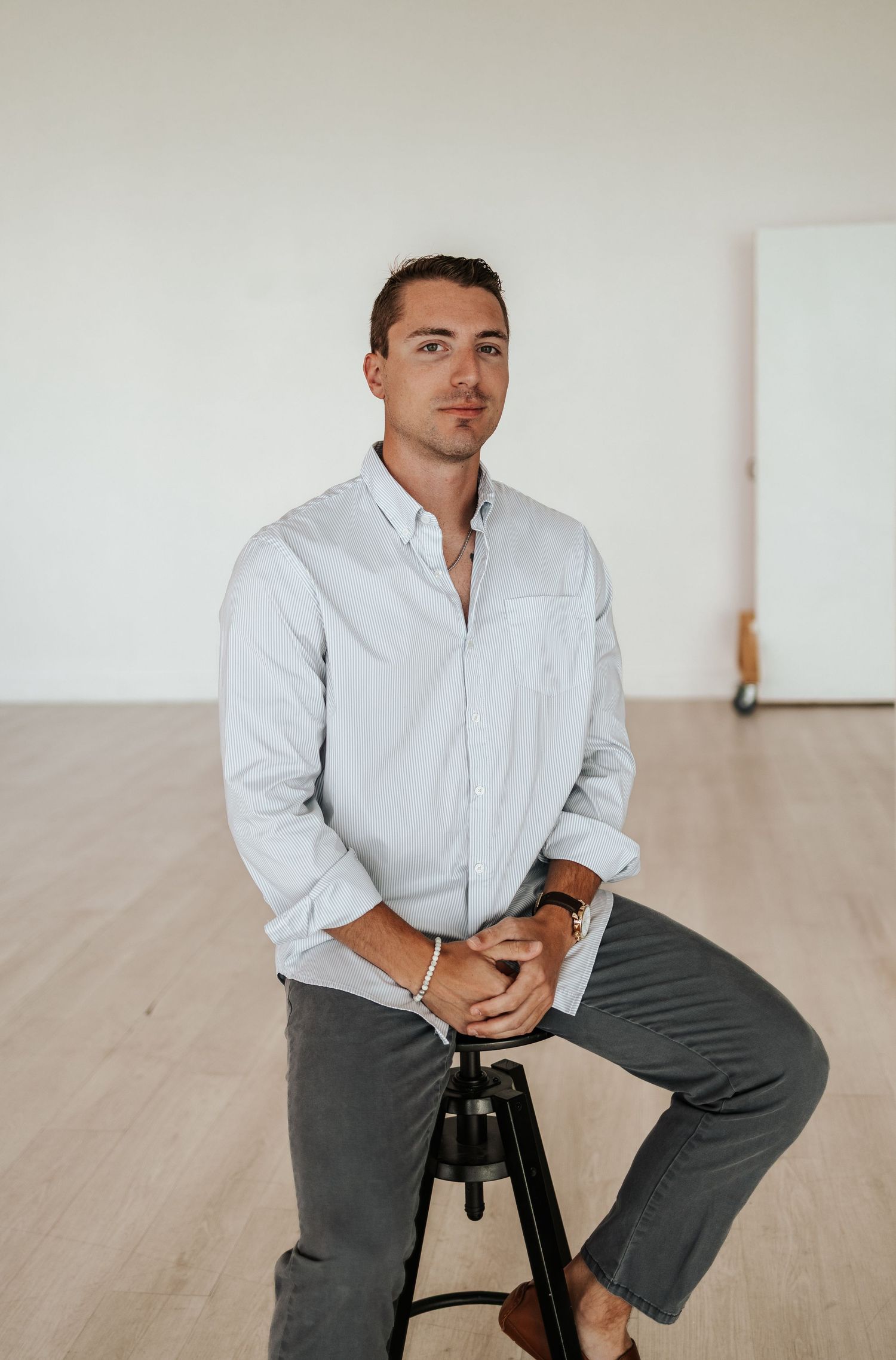 Young man in light blue button-down shirt sitting on stool in minimalist white studio space.