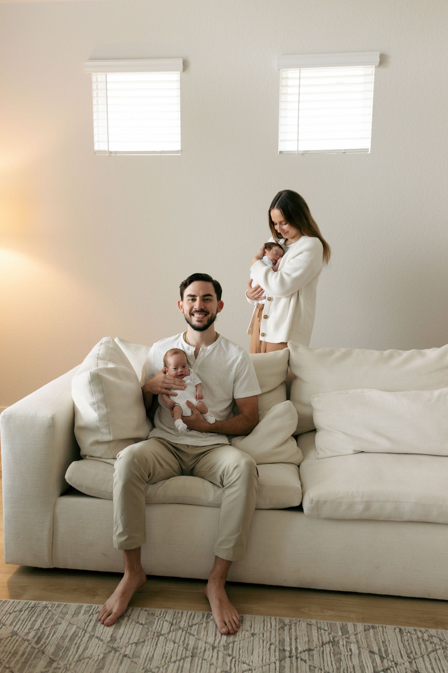 Smiling father sits on white sofa holding newborn while mother stands behind holding second twin baby.