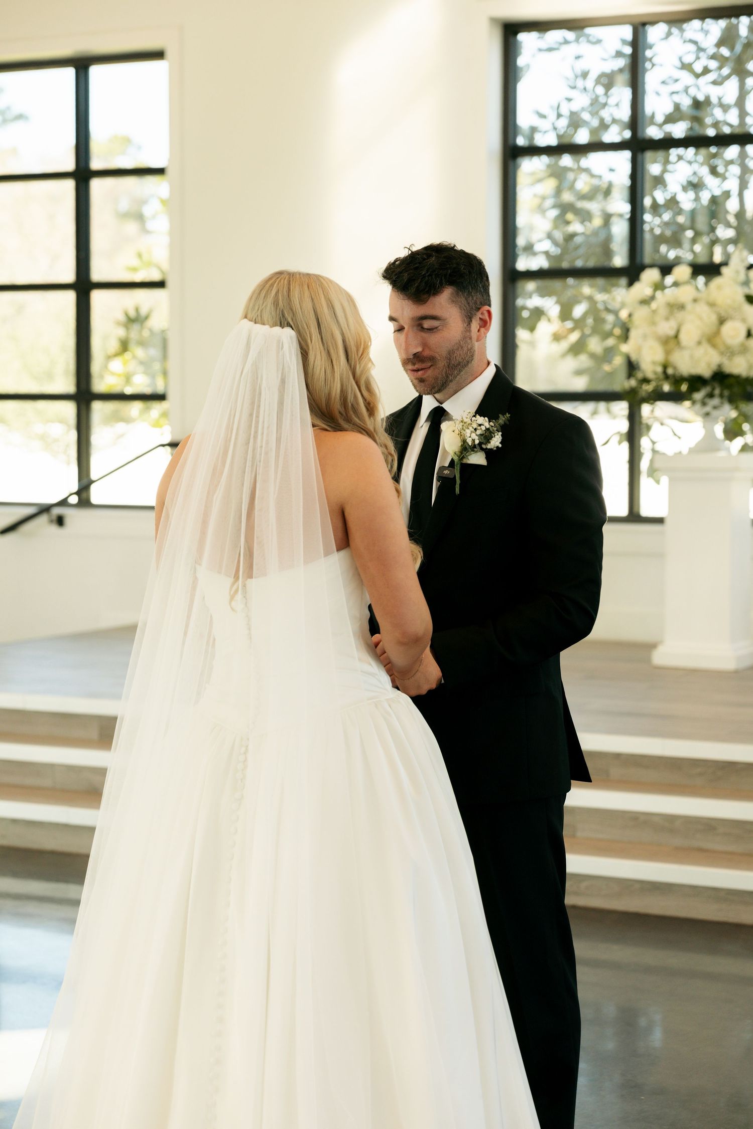 Bride in white ball gown with veil and groom in black tuxedo hold hands during indoor wedding ceremony.