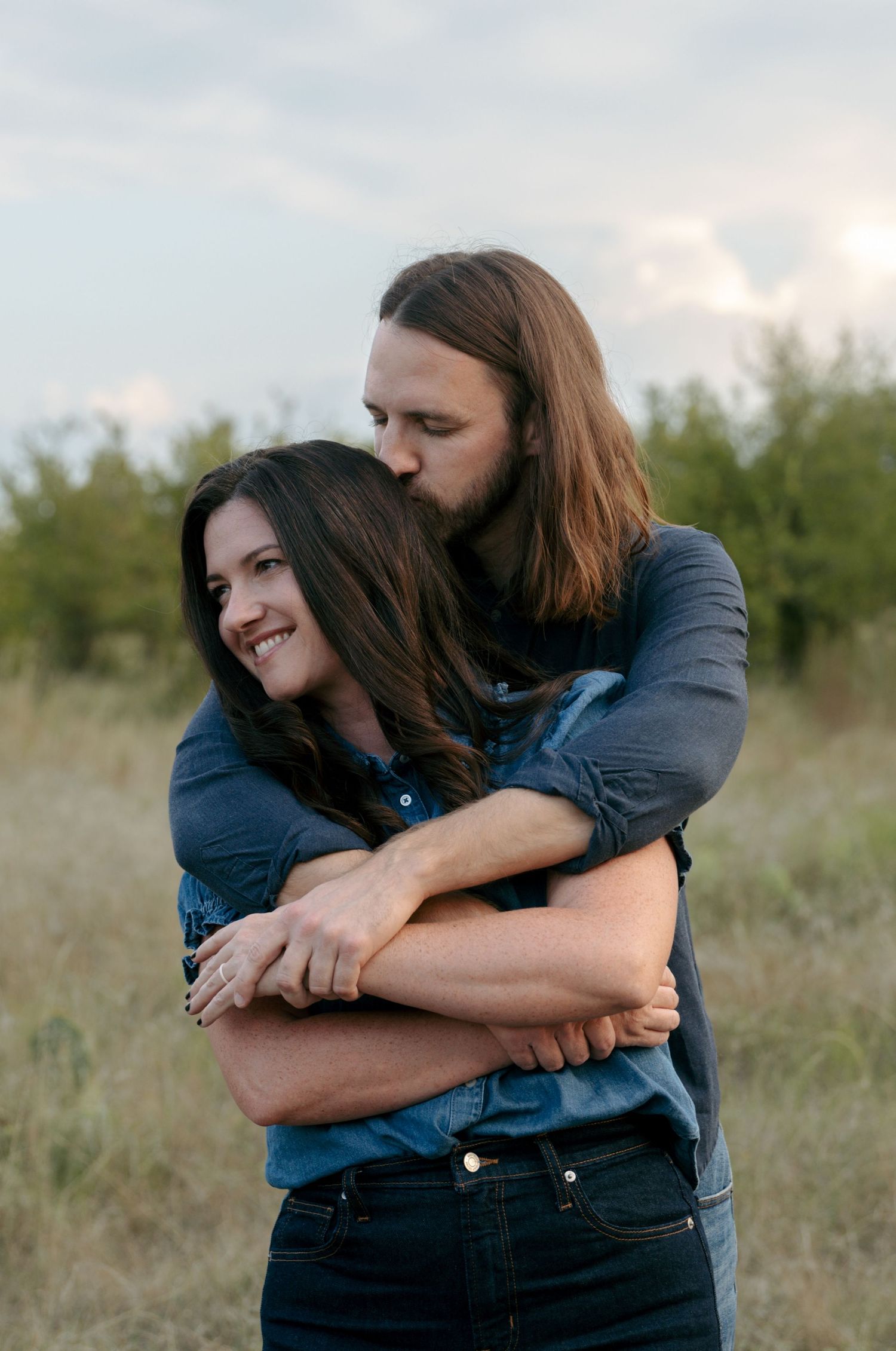Man with long hair hugging smiling woman from behind in an open grassy field.
