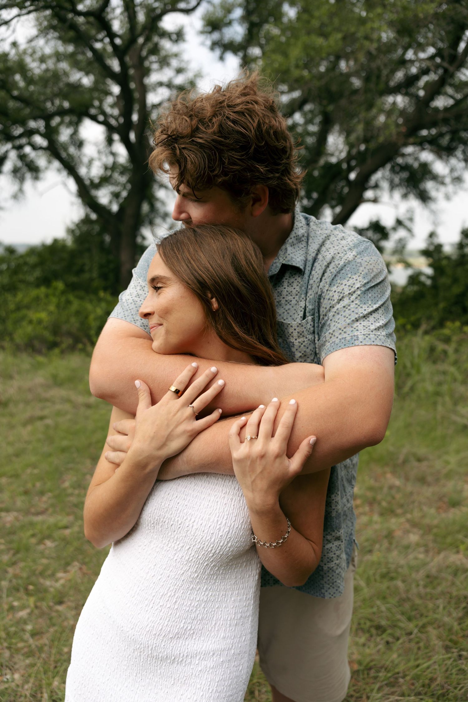 Couple embracing in a field, man hugging woman from behind in white dress near trees.