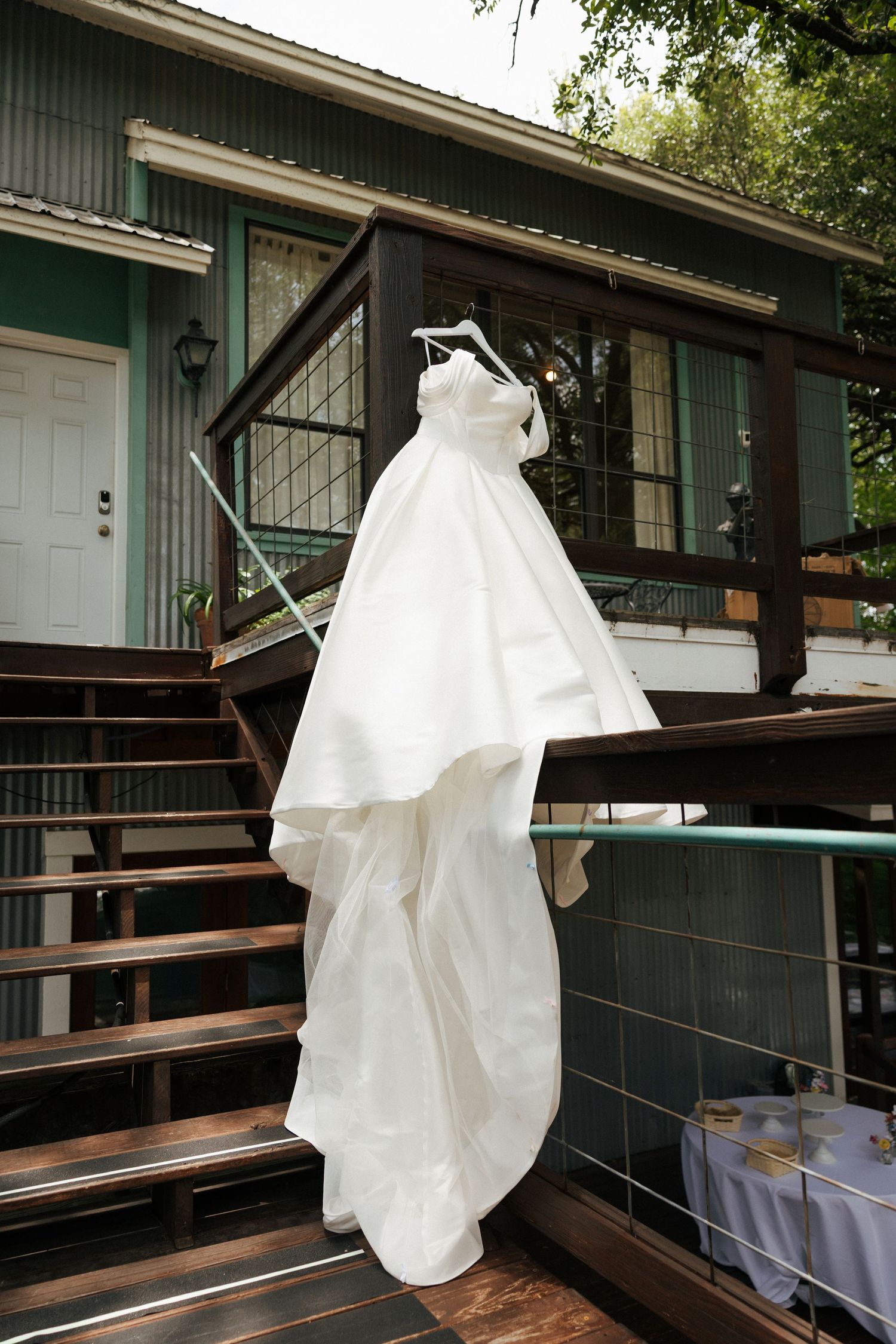 White wedding dress hanging on a hanger on a wooden deck railing outside a green house.